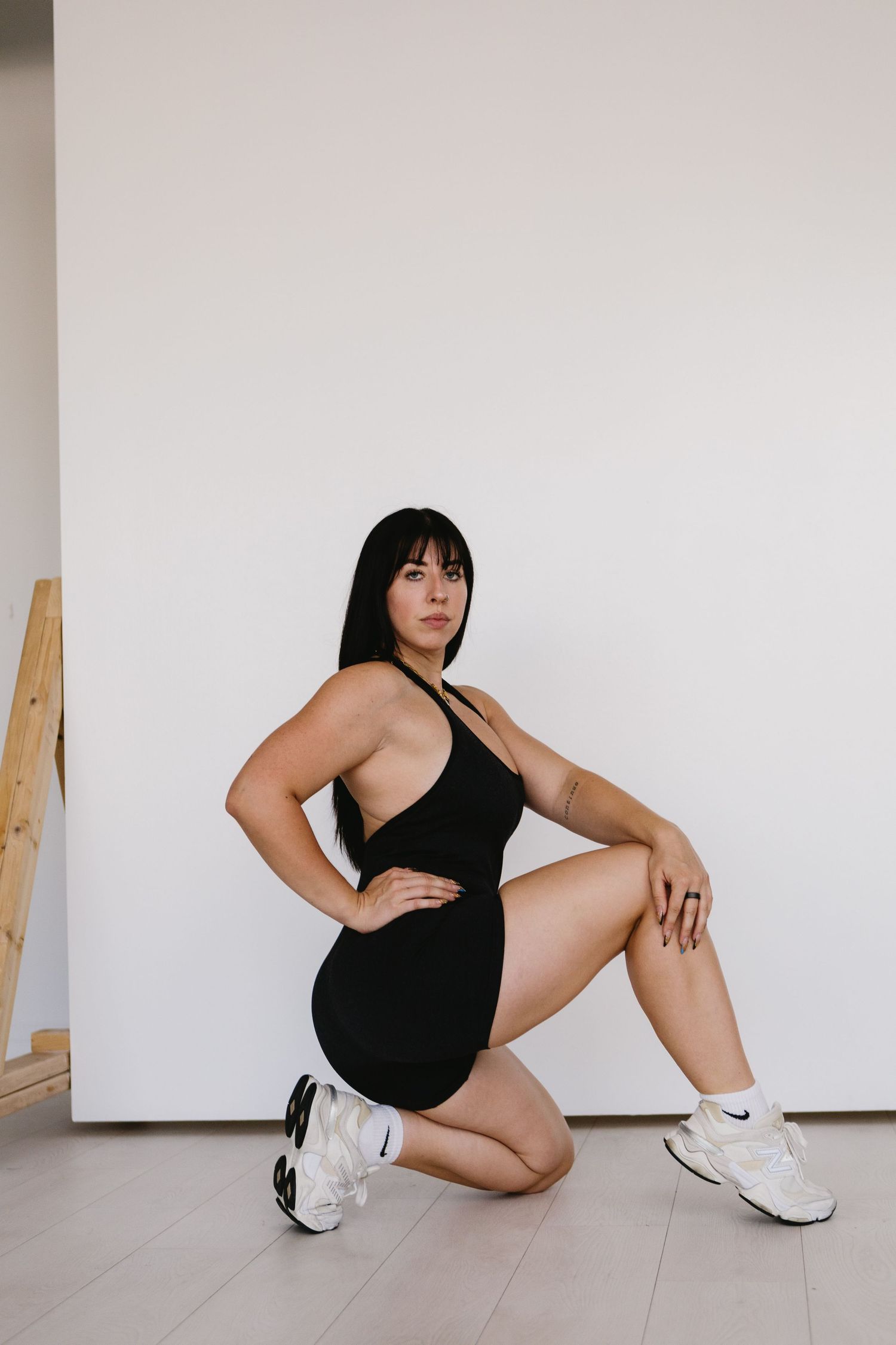 Woman in black romper and white sneakers posing in a kneeling position against a white wall.