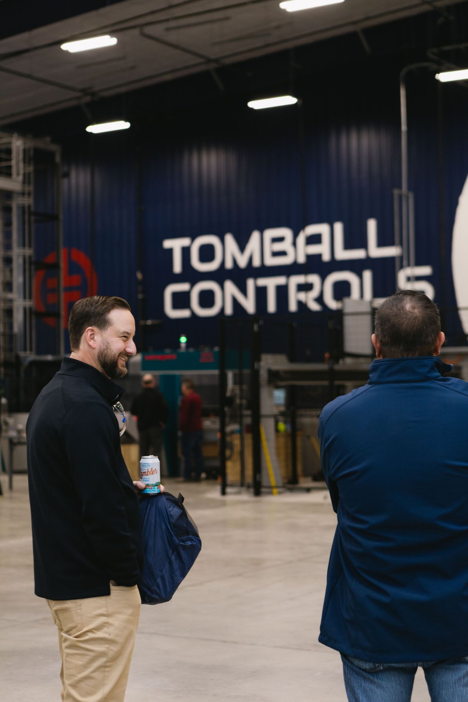 Two people having a conversation in front of a large Tomball Controls logo display in an industrial space.