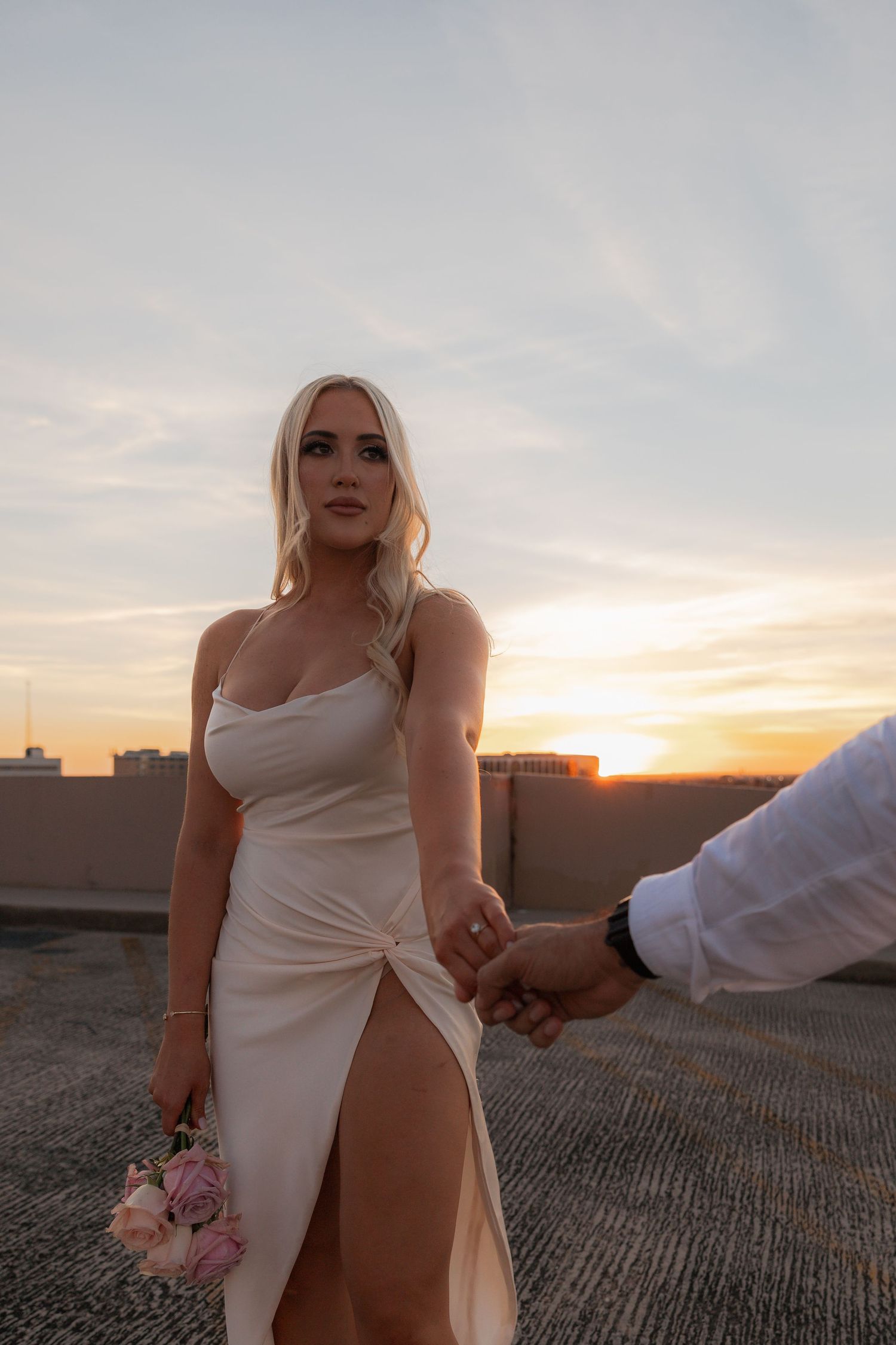 A couple in white attire poses romantically at sunset on an urban rooftop.