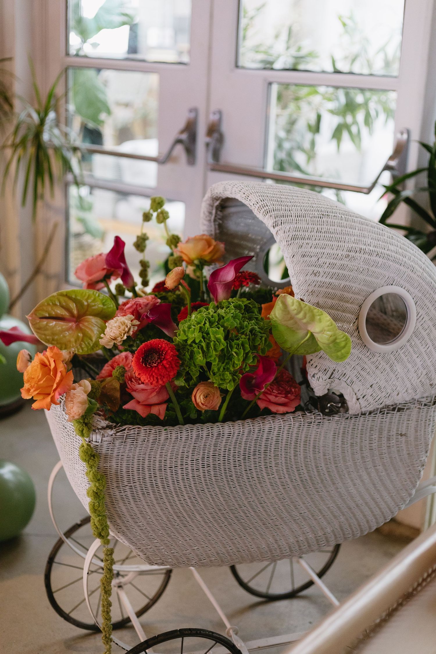 A white wicker baby carriage planter decorated with colorful fresh flowers and greenery displayed in a bright window setting.