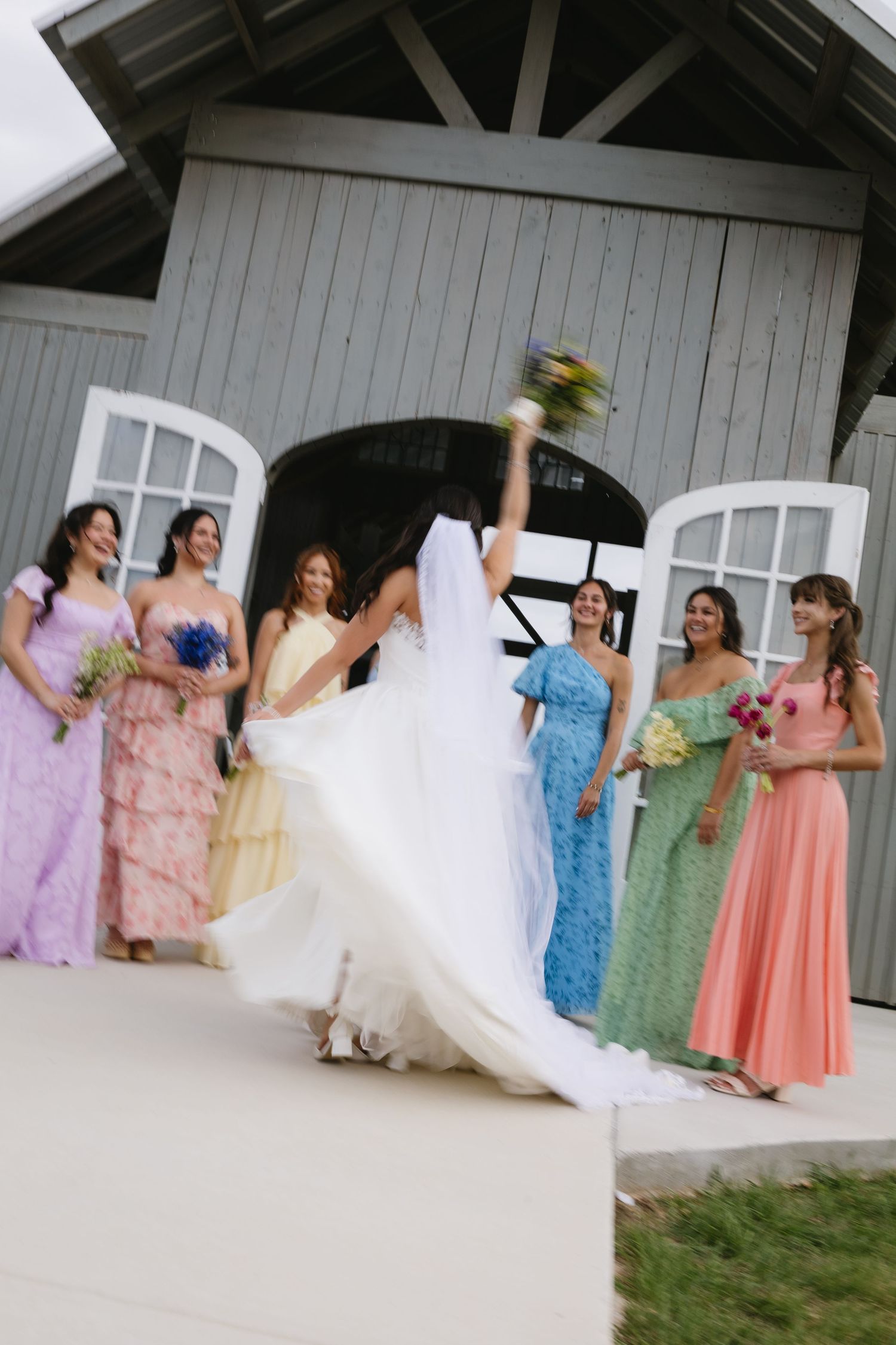 Bride in white gown raises bouquet surrounded by bridesmaids in colorful pastel dresses outside a gray barn.