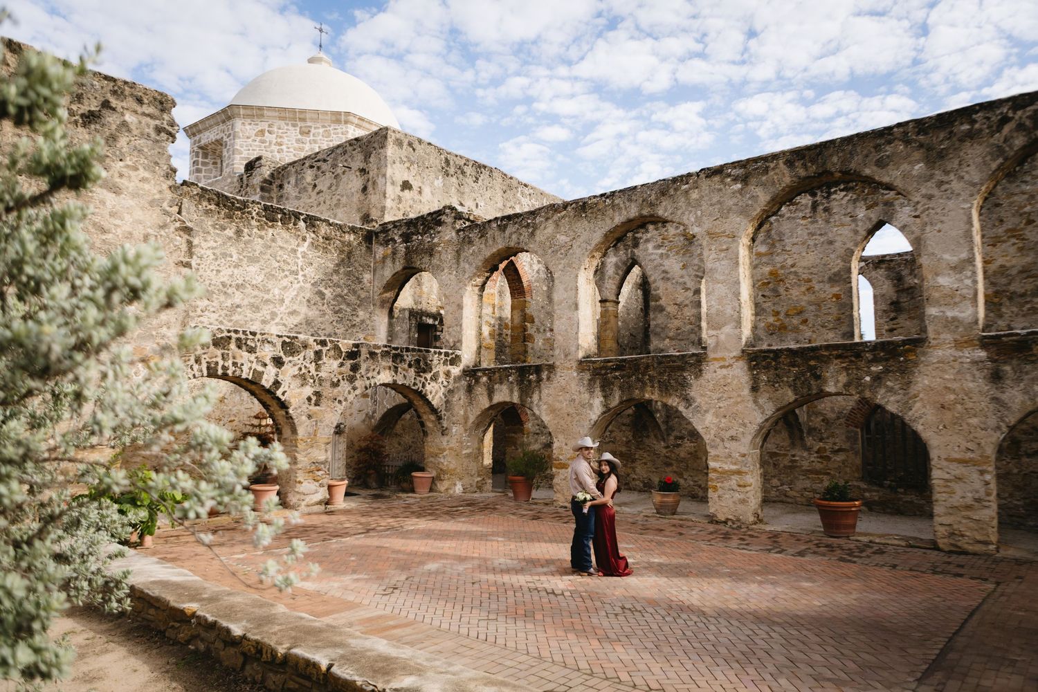 Couple embracing in historic Mission San Jose courtyard with stone arches and brick pathway in San Antonio, Texas.