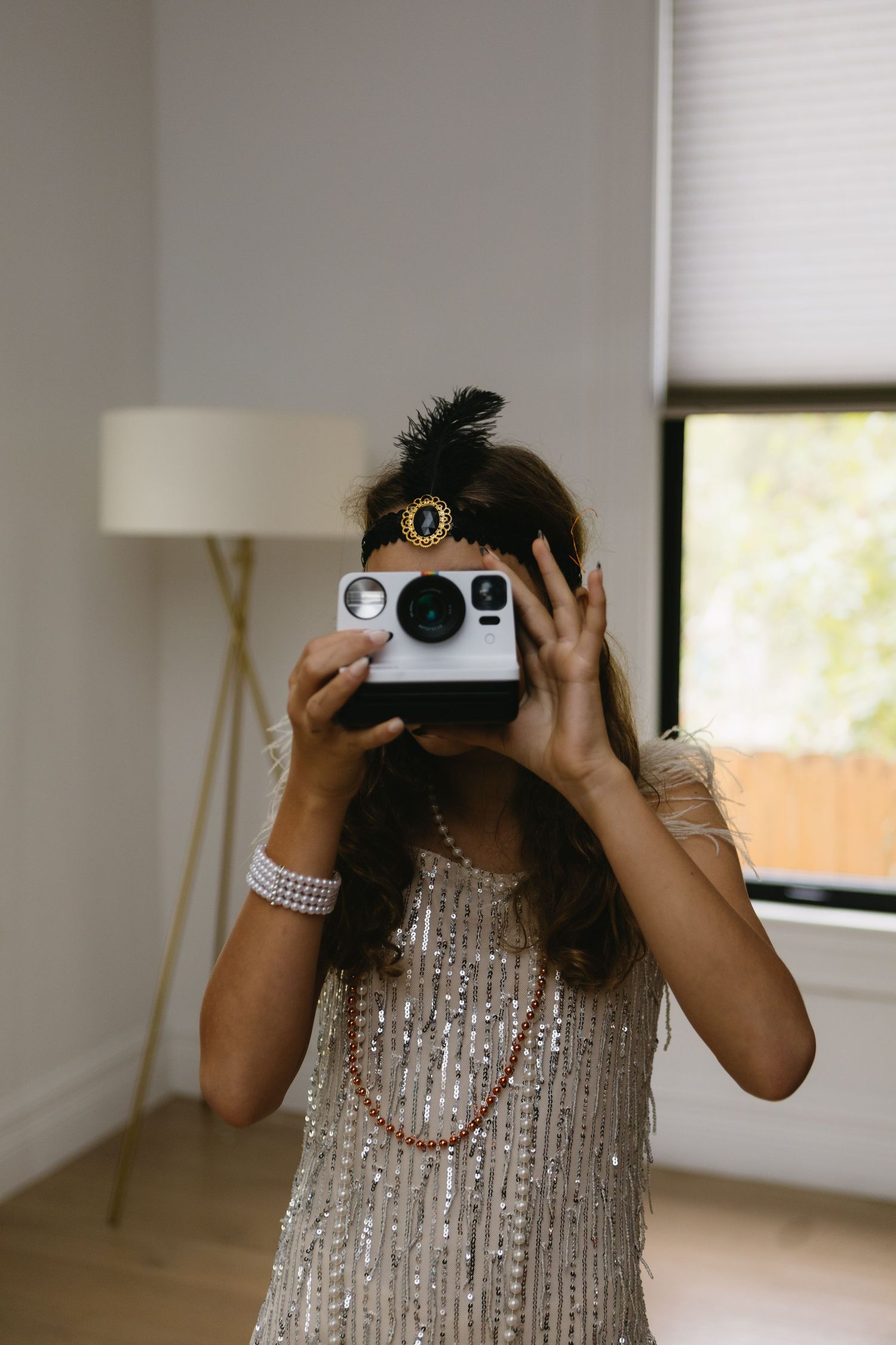 Woman in sparkly flapper dress and feather headband holds Polaroid camera up to her face indoors.