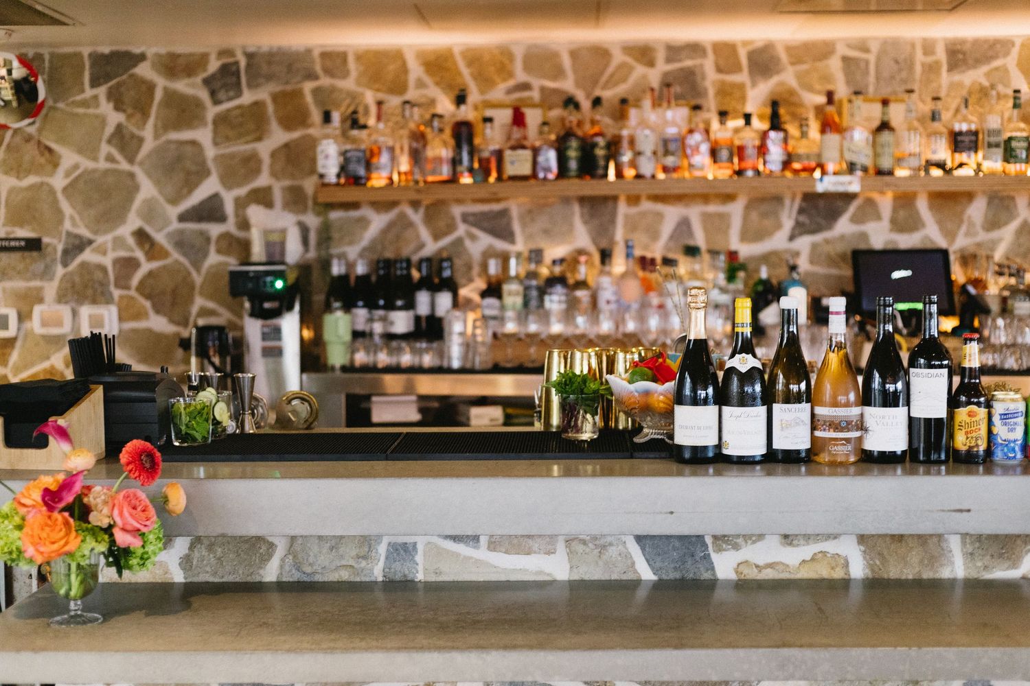 Elegant bar with stone wall backdrop, lined with wine bottles and spirits, accented by colorful fresh flowers.