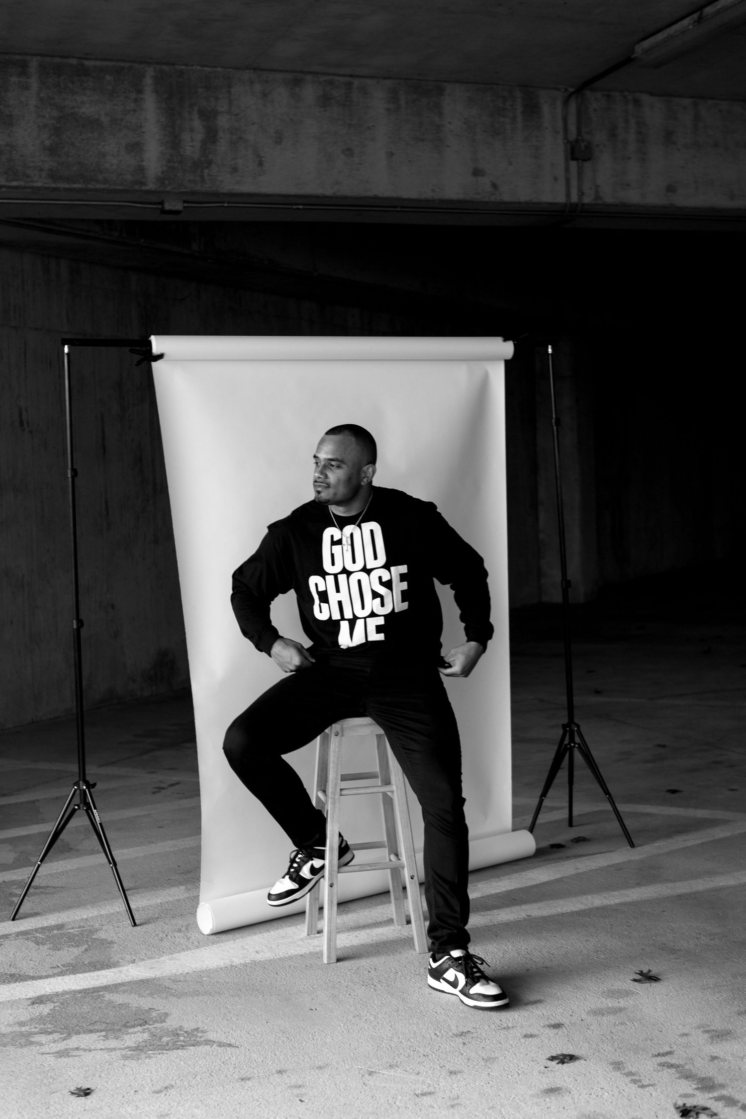Man in black God Chose Me sweatshirt poses on stool before white backdrop in black and white photo.