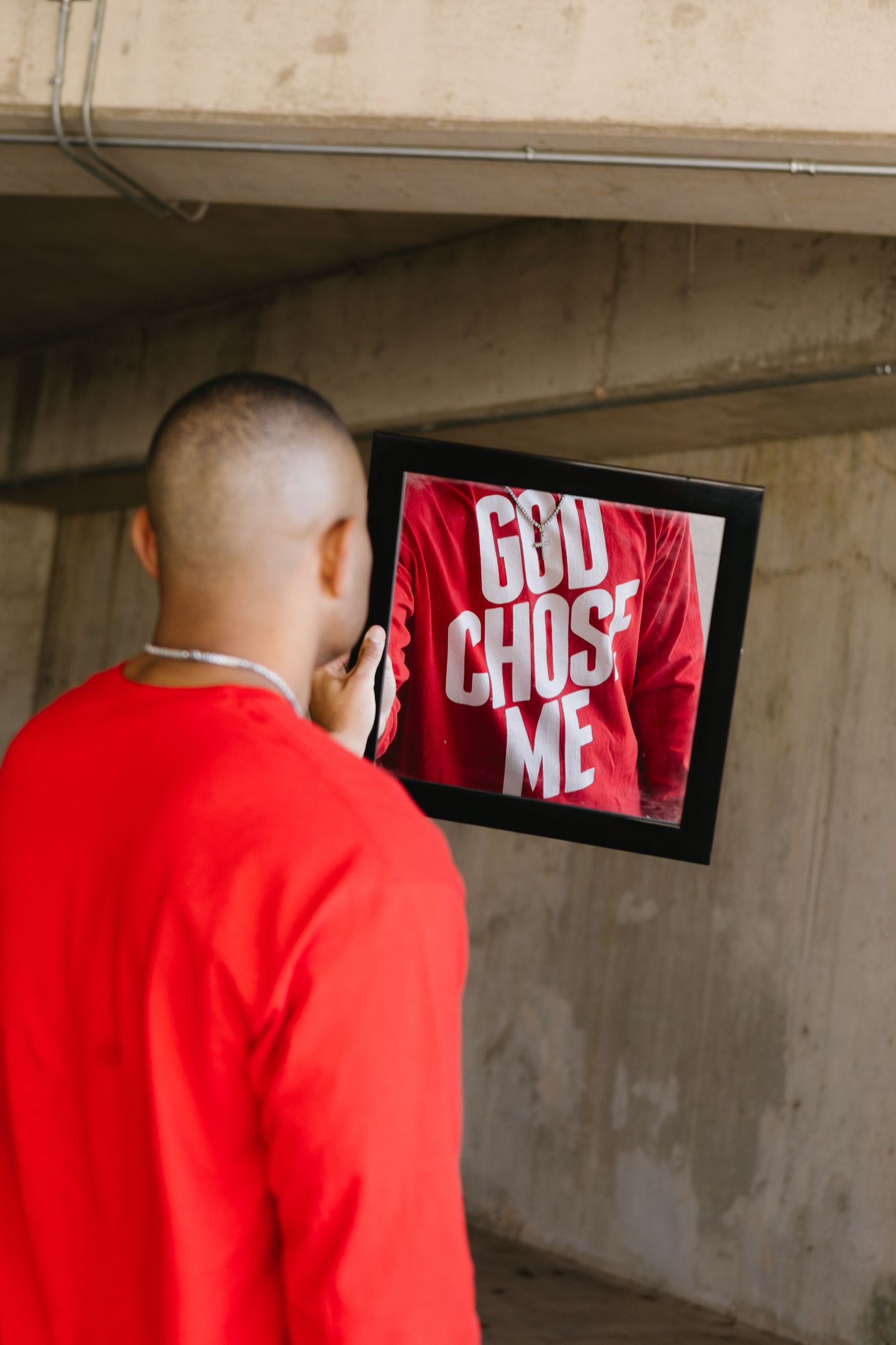 Man in red sweatshirt holding framed photo showing GOD CHOSE ME shirt against concrete wall.