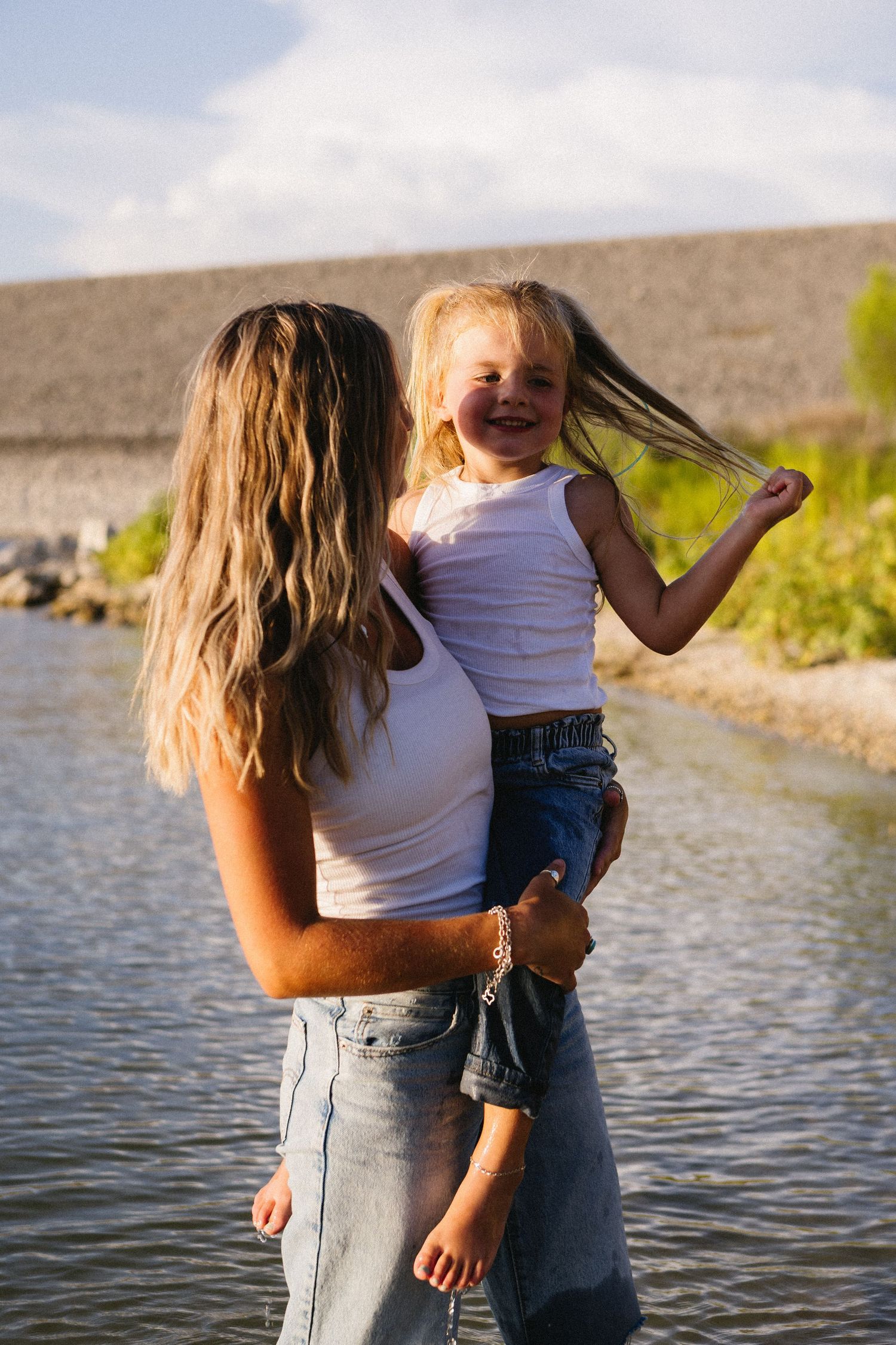 A woman holding a laughing blonde toddler near a lake, both wearing white tank tops and jeans outdoors.