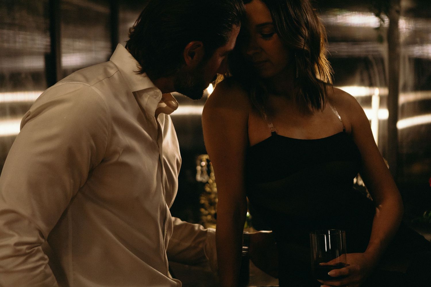 A man in a white shirt leans close to a woman in a black dress holding a wine glass in a dimly lit bar.