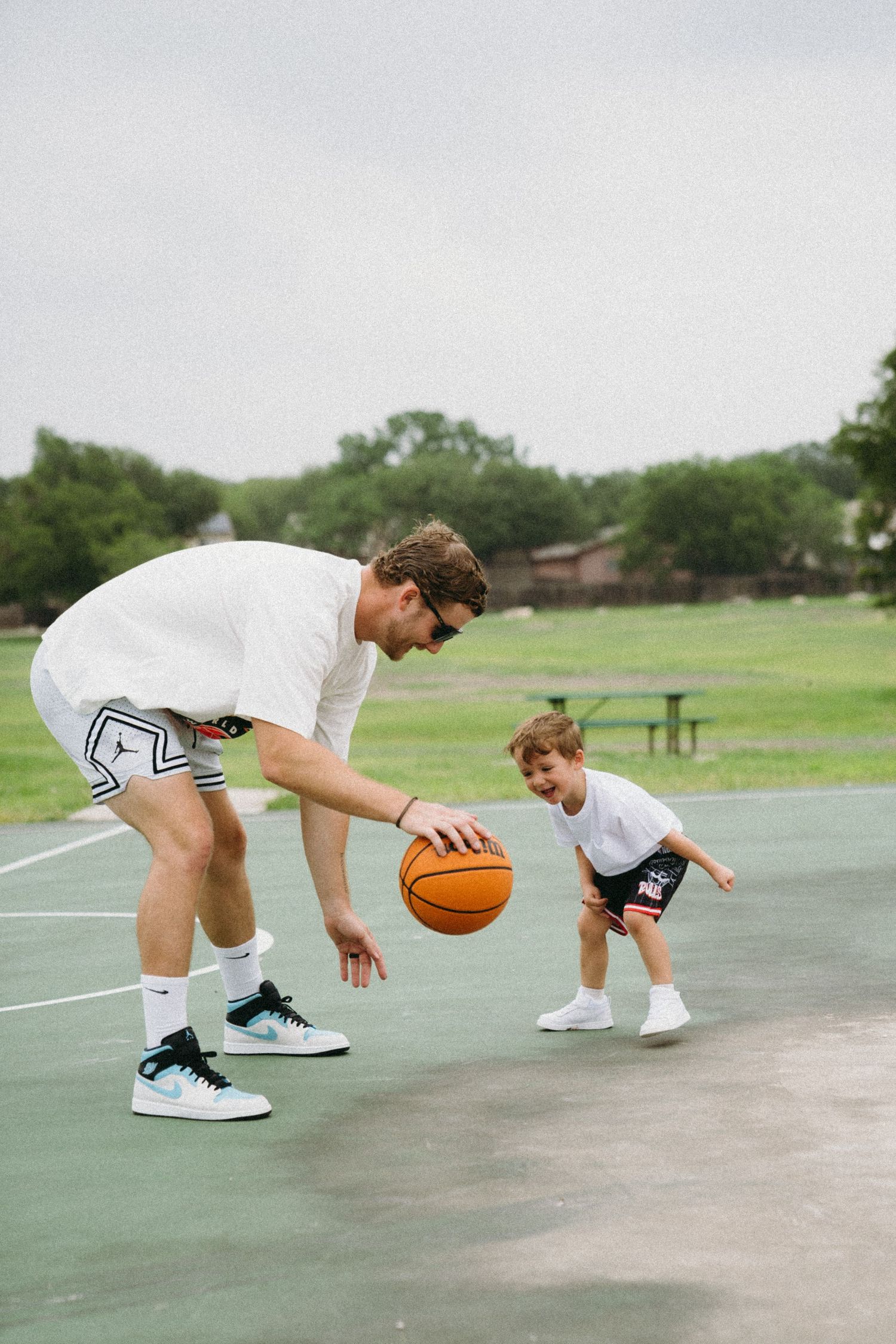 Adult and child playing basketball together on an outdoor court, dribbling an orange ball on a cloudy day.
