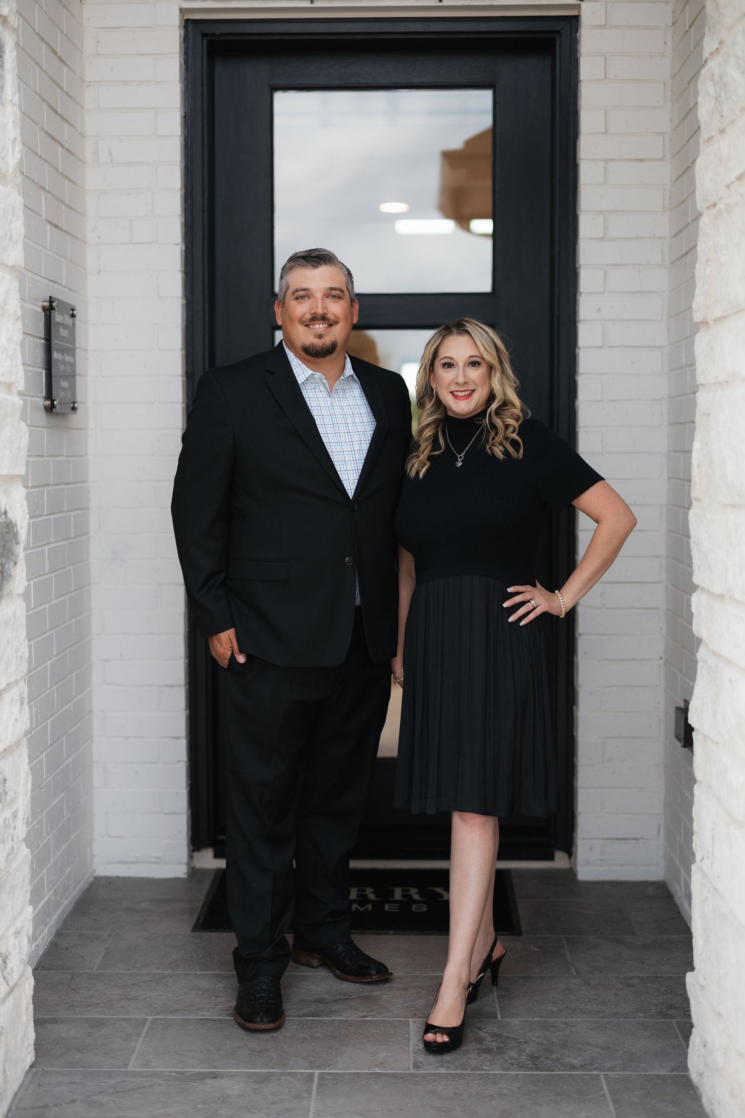 A group in formal black attire poses in front of white brick walls and dark doorways during a photo shoot.