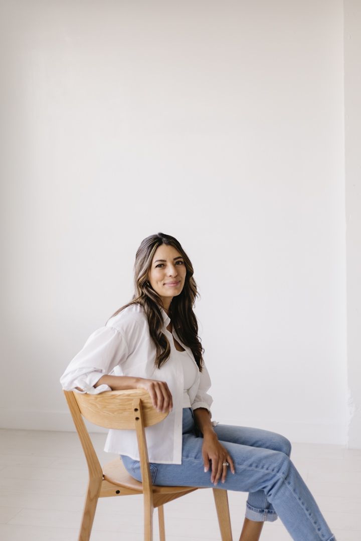 A person in a white button-down shirt and jeans sits casually on a wooden chair against a minimalist white background.