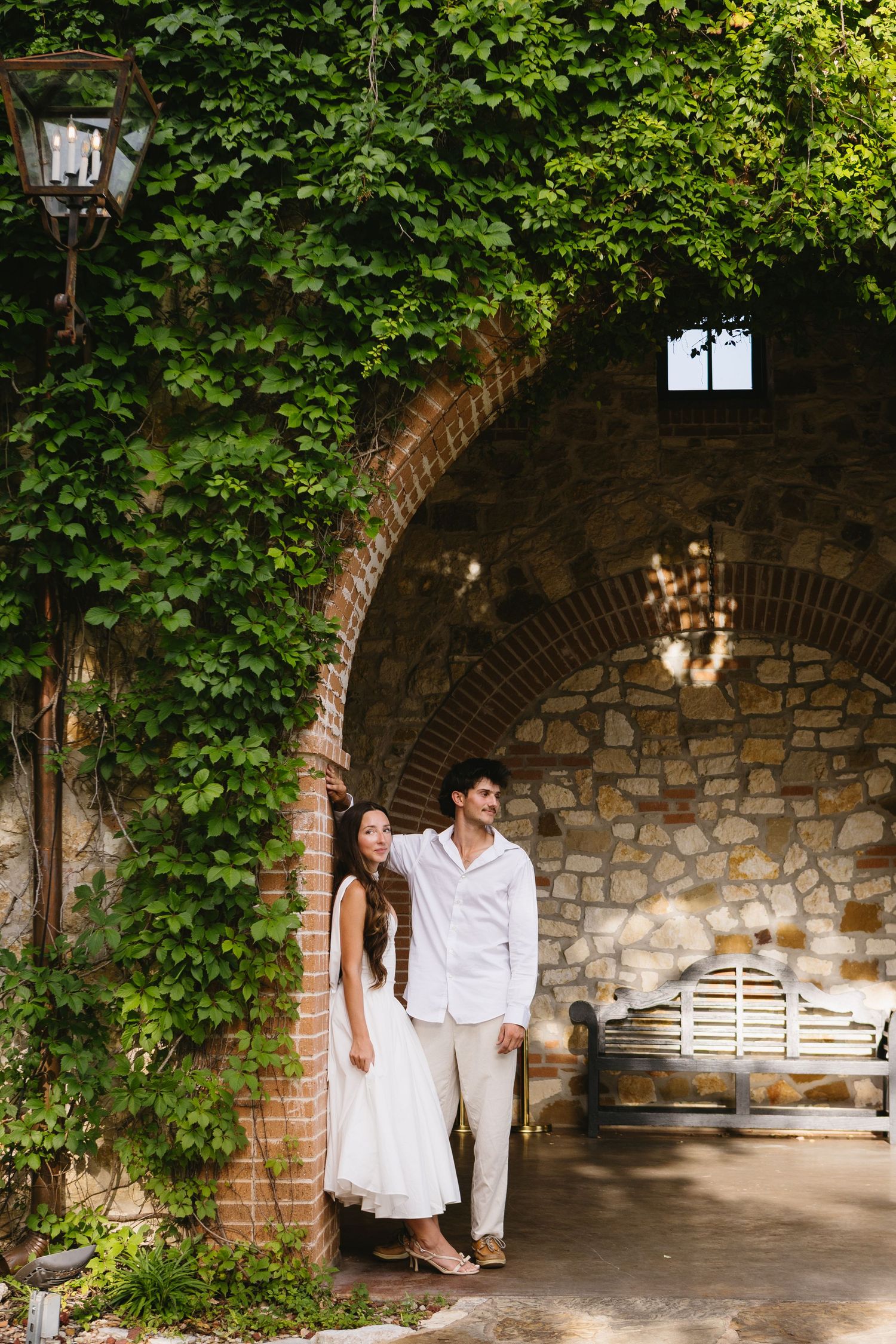 Couple in white attire posing under ivy-covered brick arch at rustic stone villa, romantic outdoor setting.