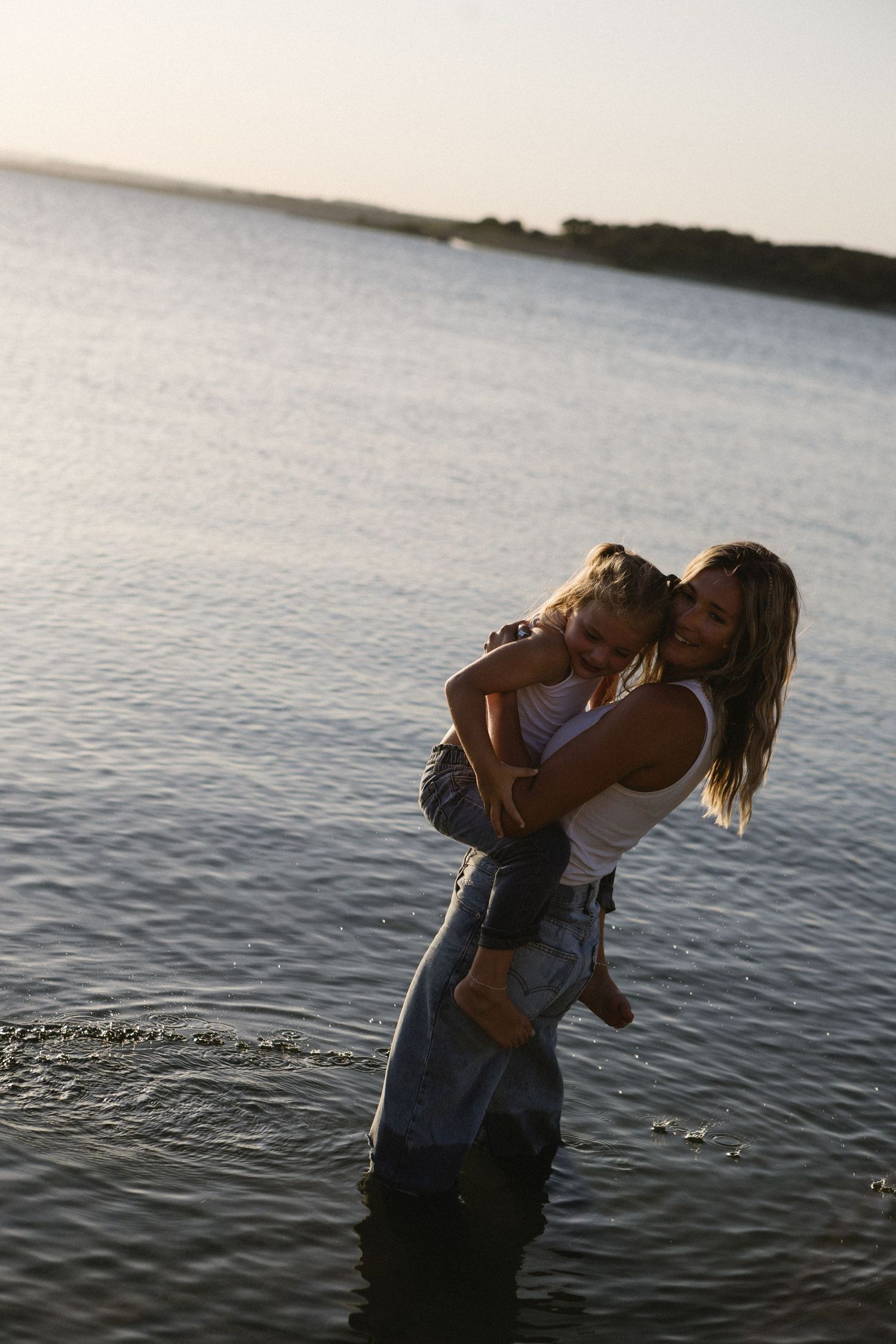 Woman holding a young child while standing in calm water at sunset, sharing a tender moment.