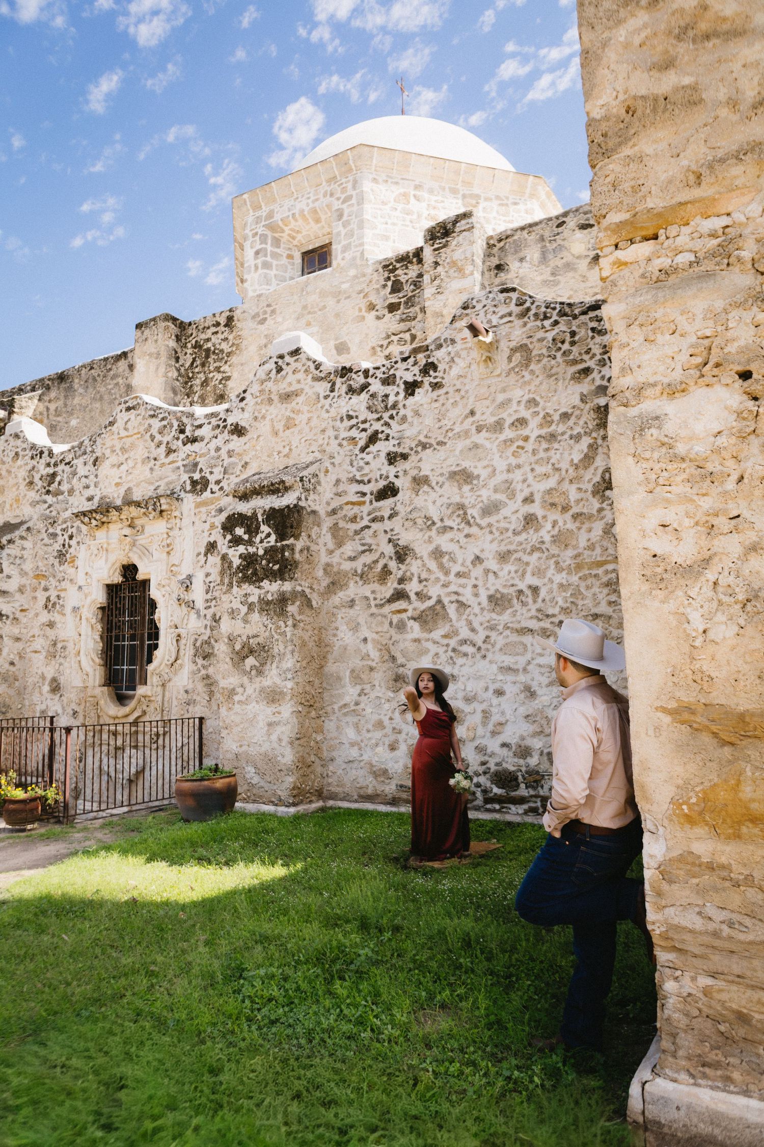 Tourists photographing historic stone mission ruins with dome architecture under blue sky in San Antonio, Texas.