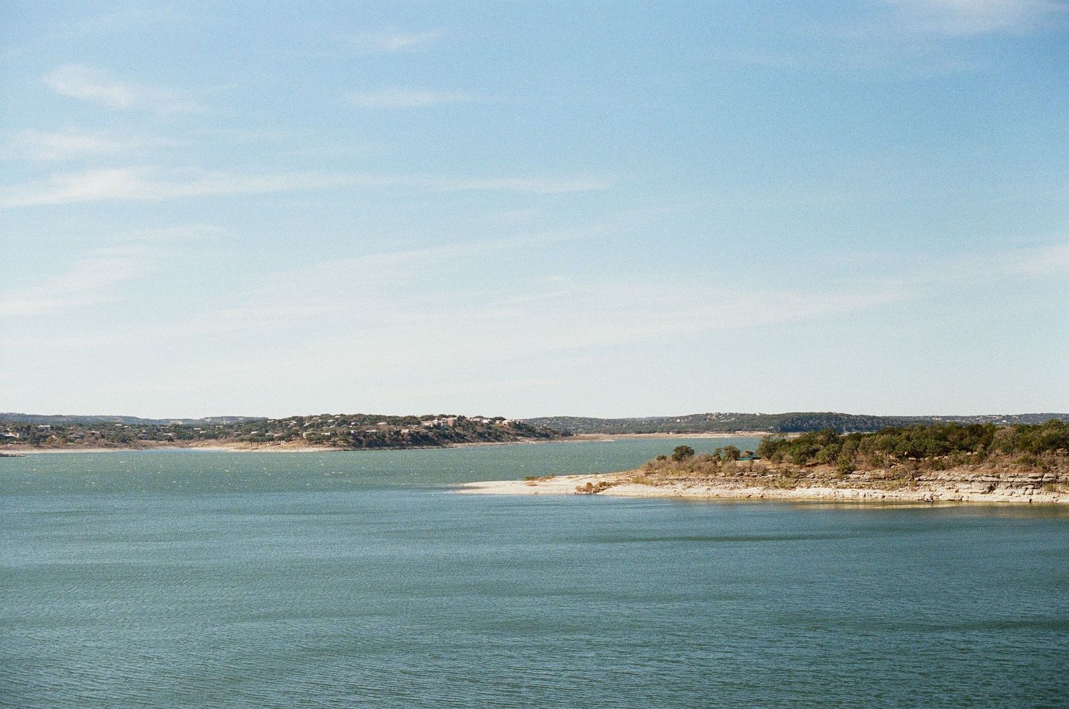 Scenic lake view with calm blue waters, rocky shoreline, and distant hills under a partly cloudy sky.