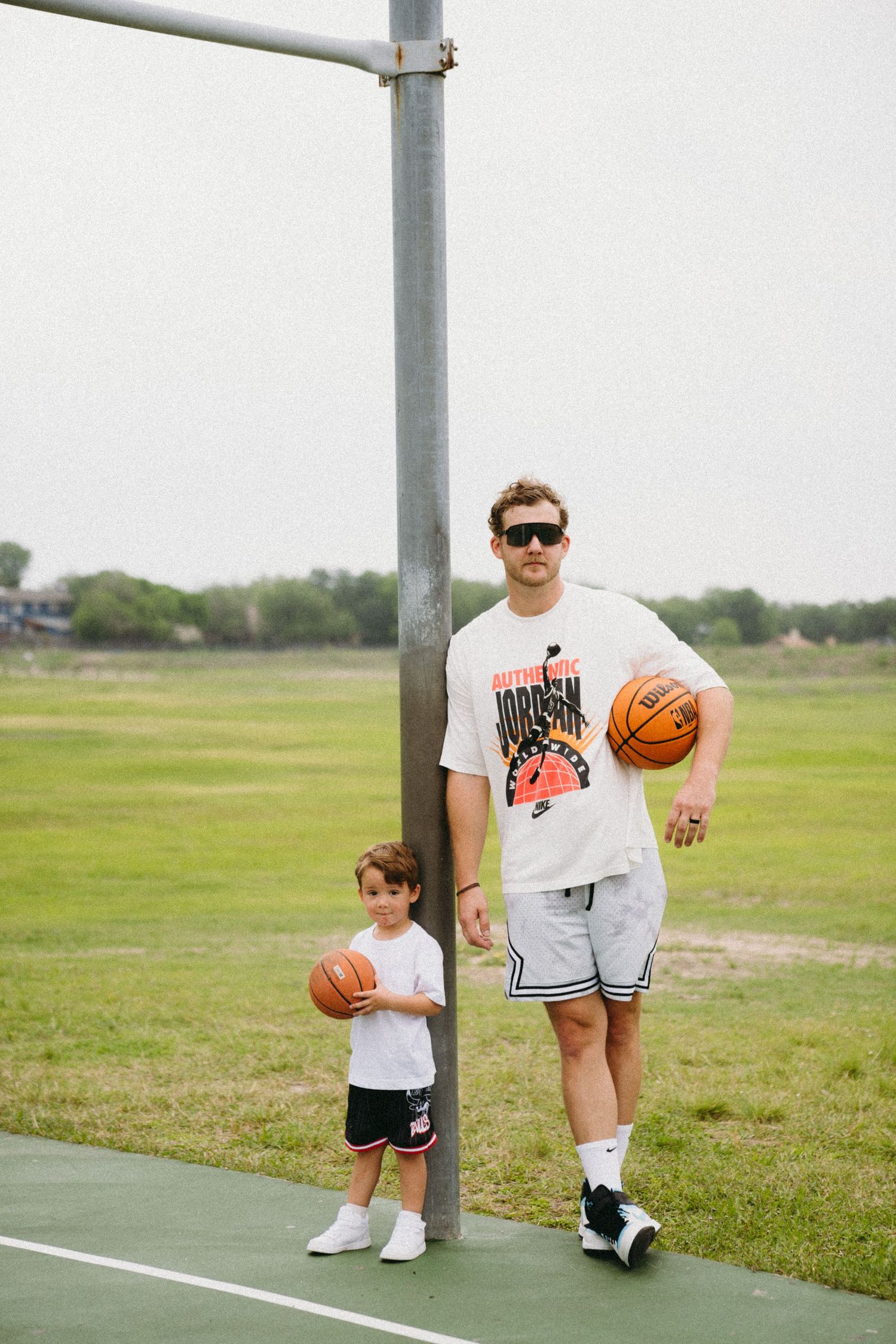 Man and young boy holding basketballs on outdoor court, wearing matching Nike Jordan outfits.