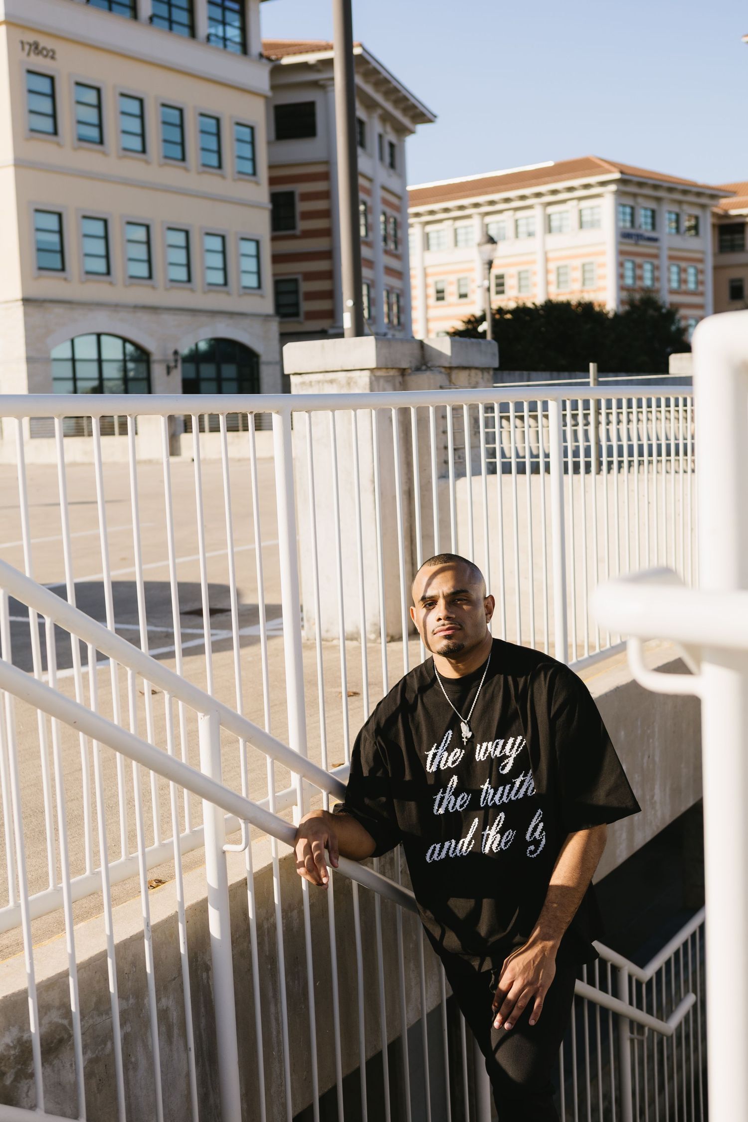 Man in black graphic tee stands on outdoor staircase with white railings and urban buildings in background.