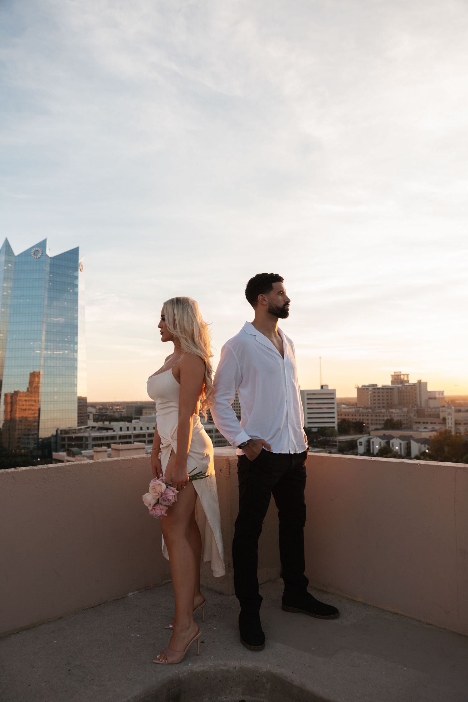 A couple stands together on an urban rooftop with glass buildings in the background.