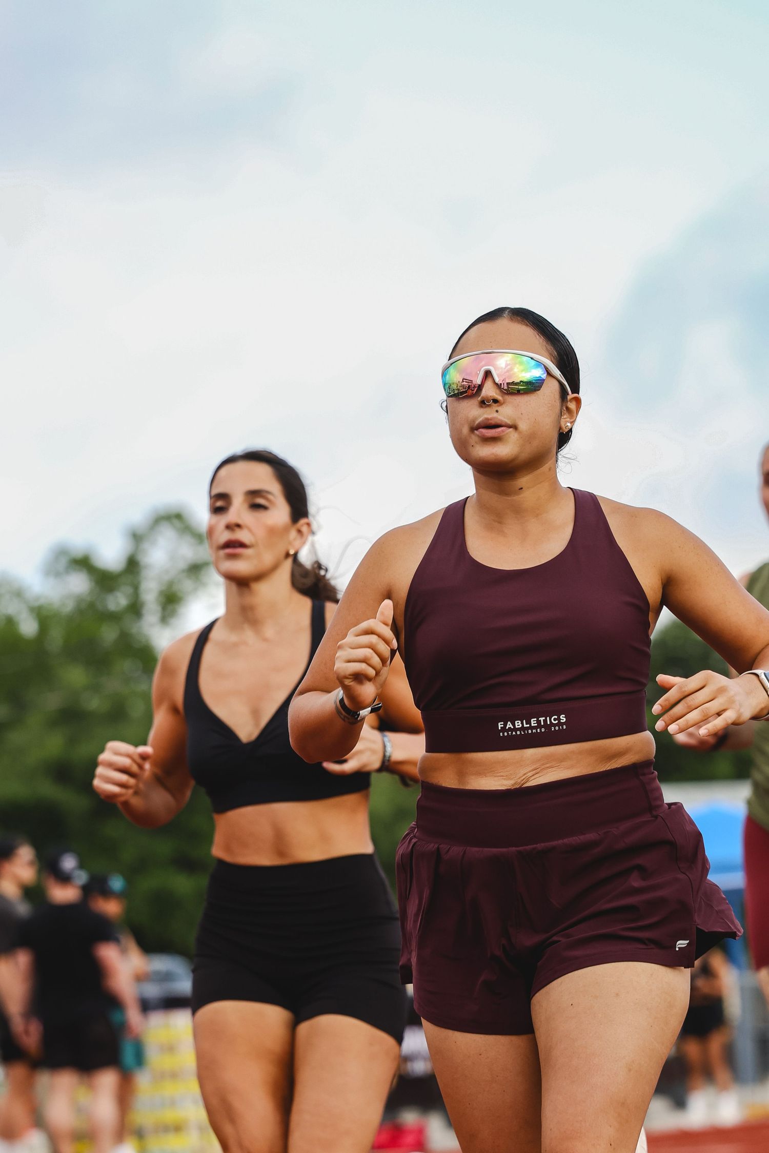 Two athletic women in sportswear running on a track, one wearing rainbow sunglasses and a maroon Fabletics outfit.