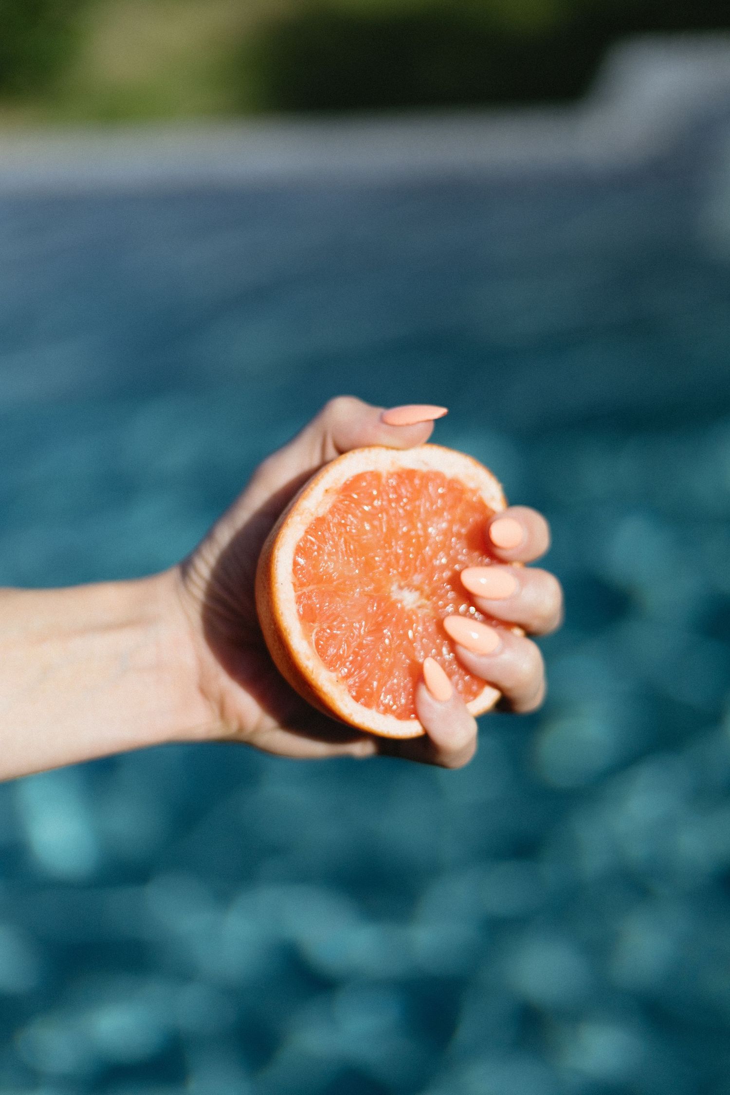 Hand holding a fresh halved grapefruit over a turquoise swimming pool on a sunny day.