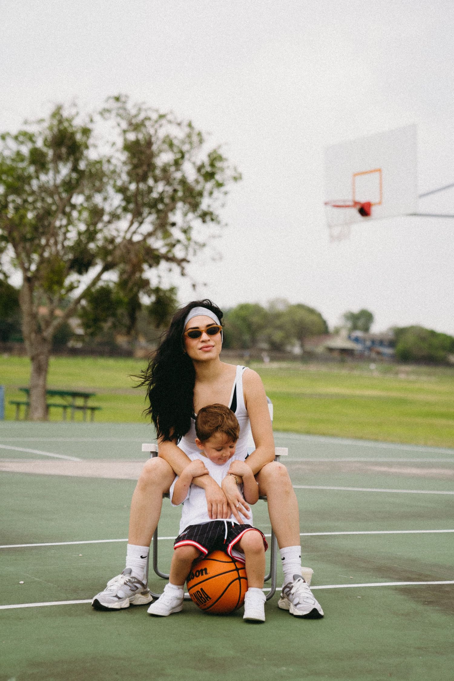 Woman in white tank top sits on chair with toddler on outdoor basketball court, basketball nearby, hoop visible in background.