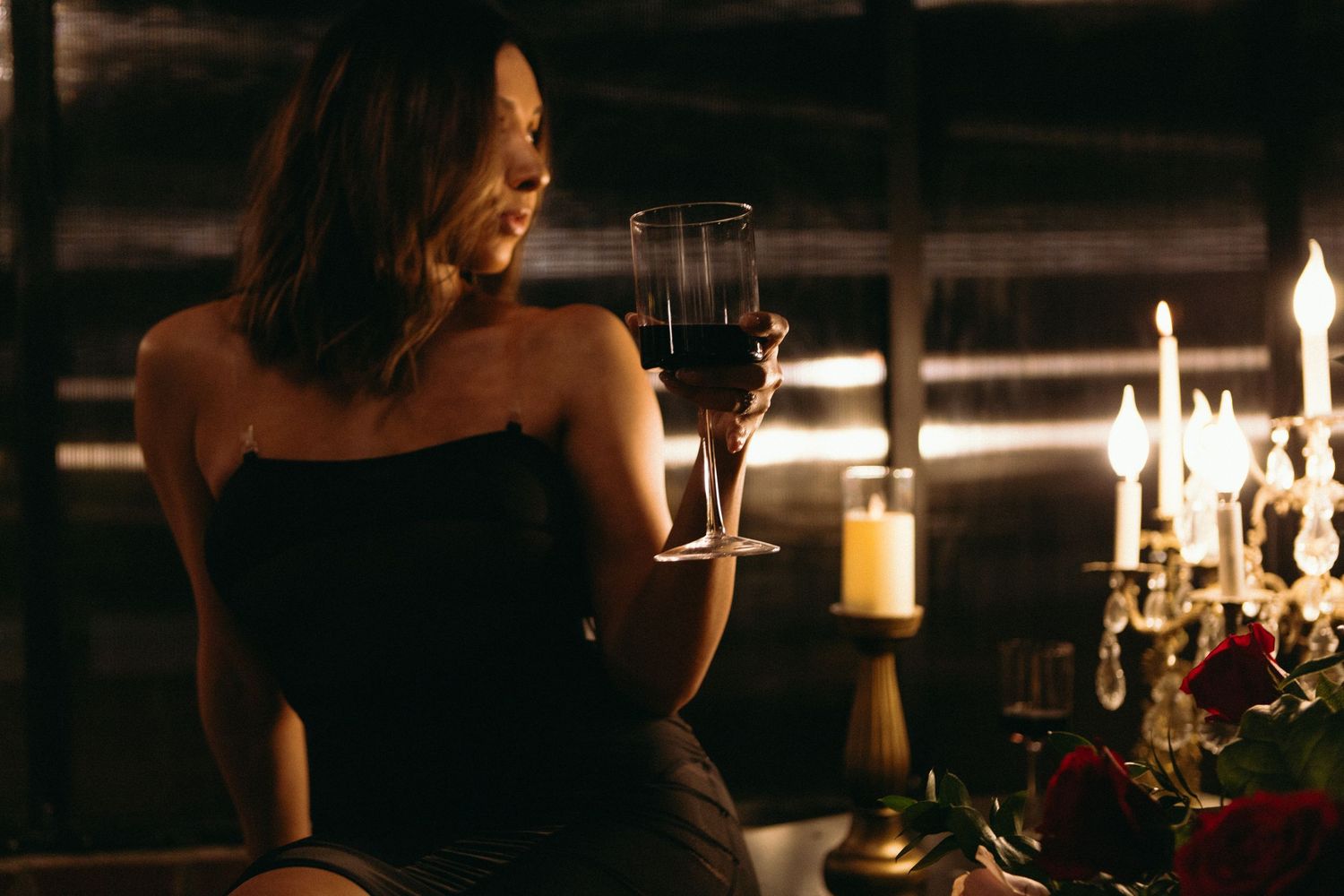 Woman in black dress holding red wine glass by candlelight at romantic dinner setting with roses.