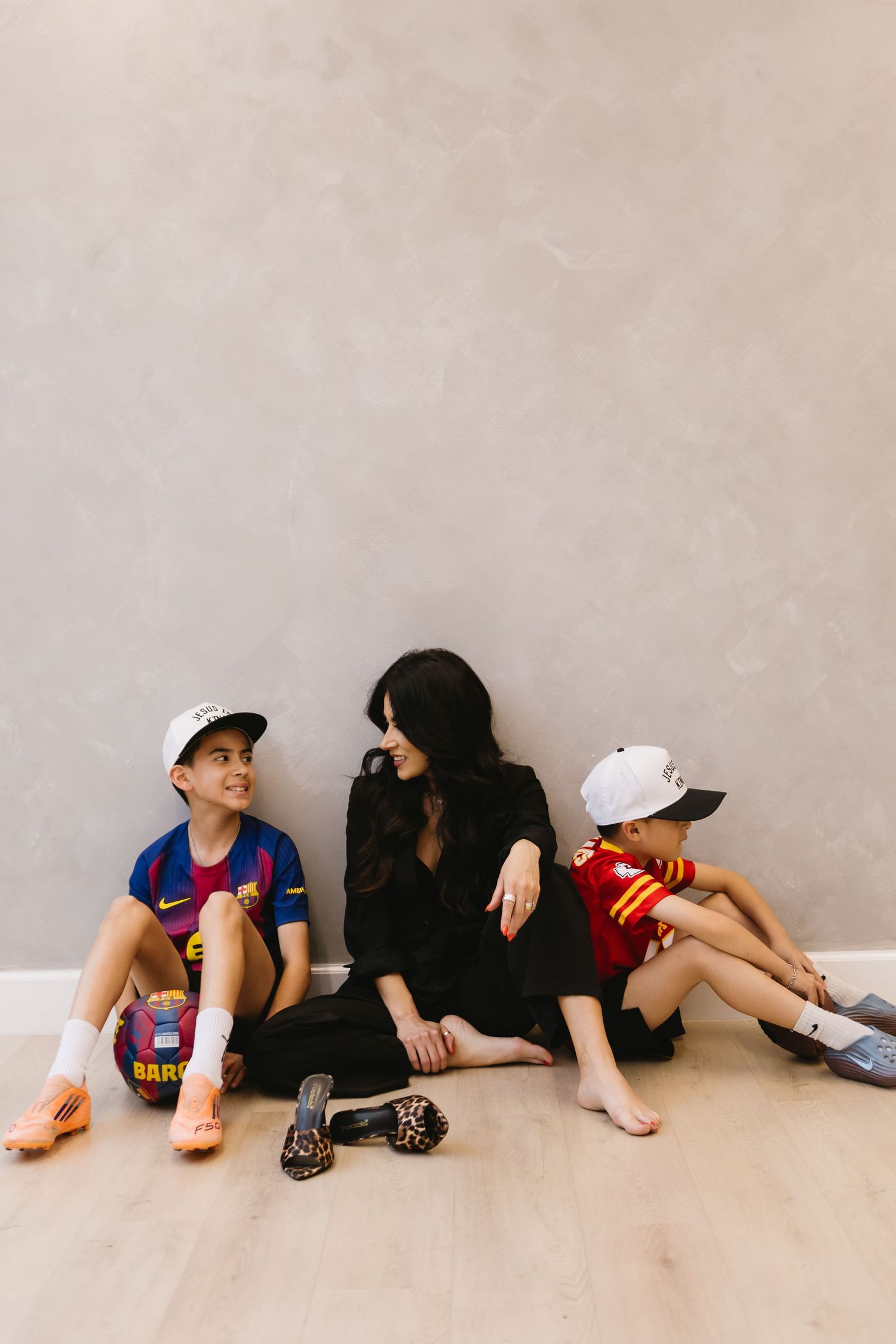 Woman in black dress sits on floor with two children in sports jerseys and caps against a gray wall.