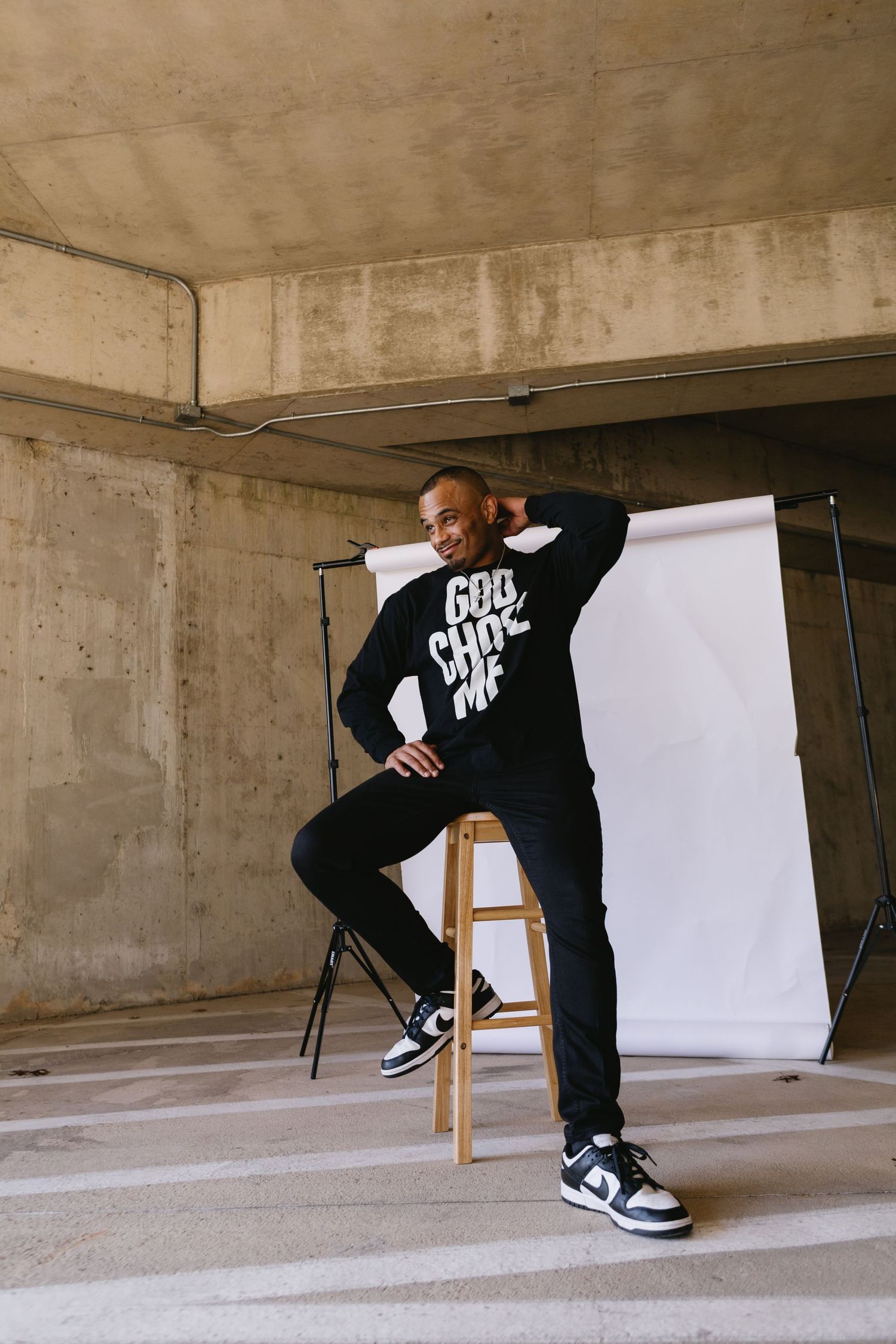 Man in black God Choose Me hoodie posing on stool in urban concrete setting with photography backdrop.