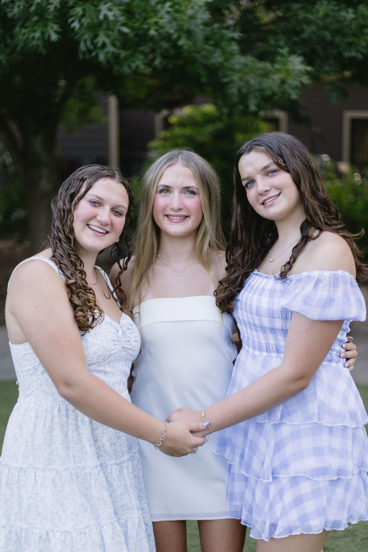 Three teenage girls in summer dresses smile and hold hands outdoors in a garden setting.