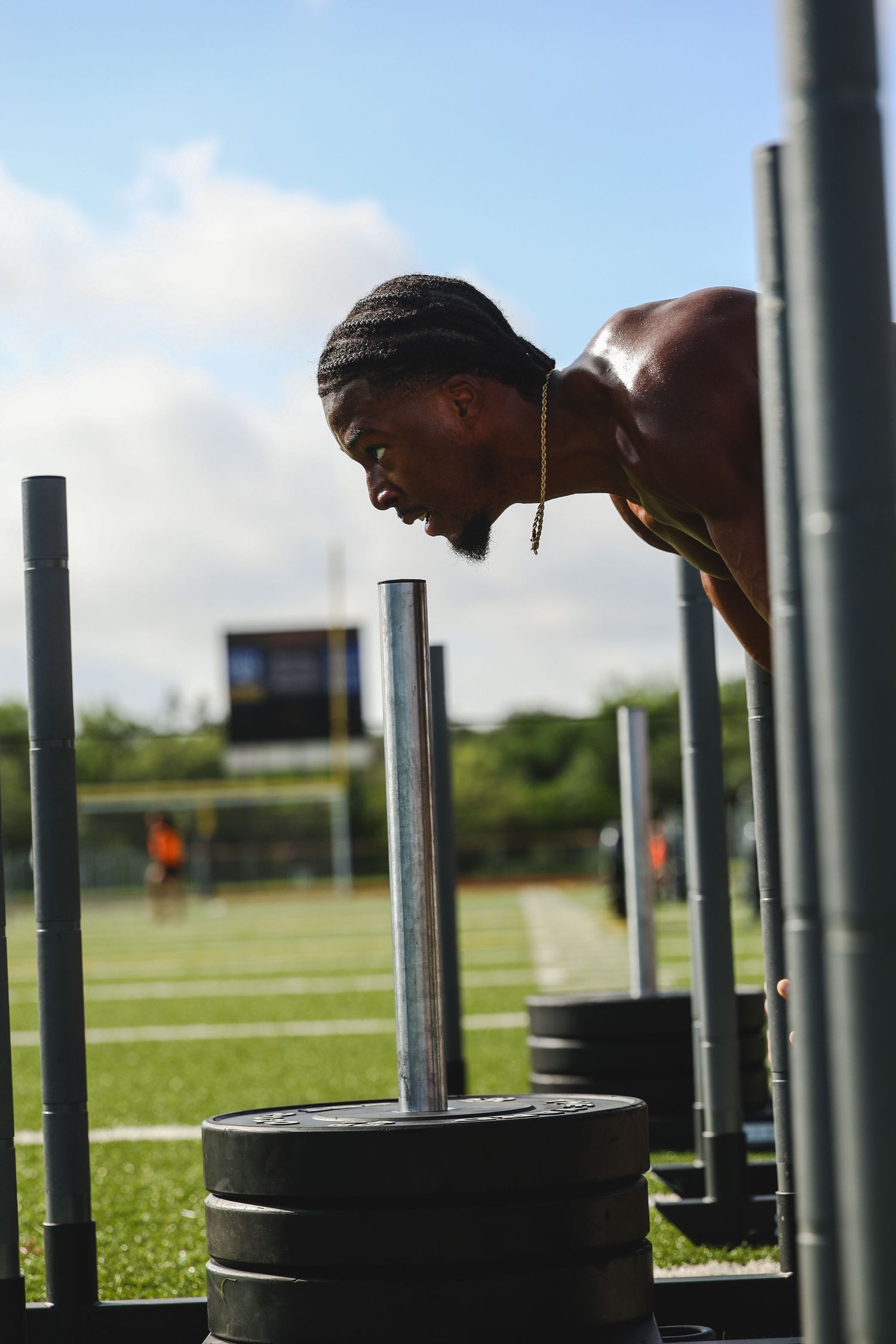 Muscular shirtless man intensely pushing heavy weighted sled on green turf field outdoors.