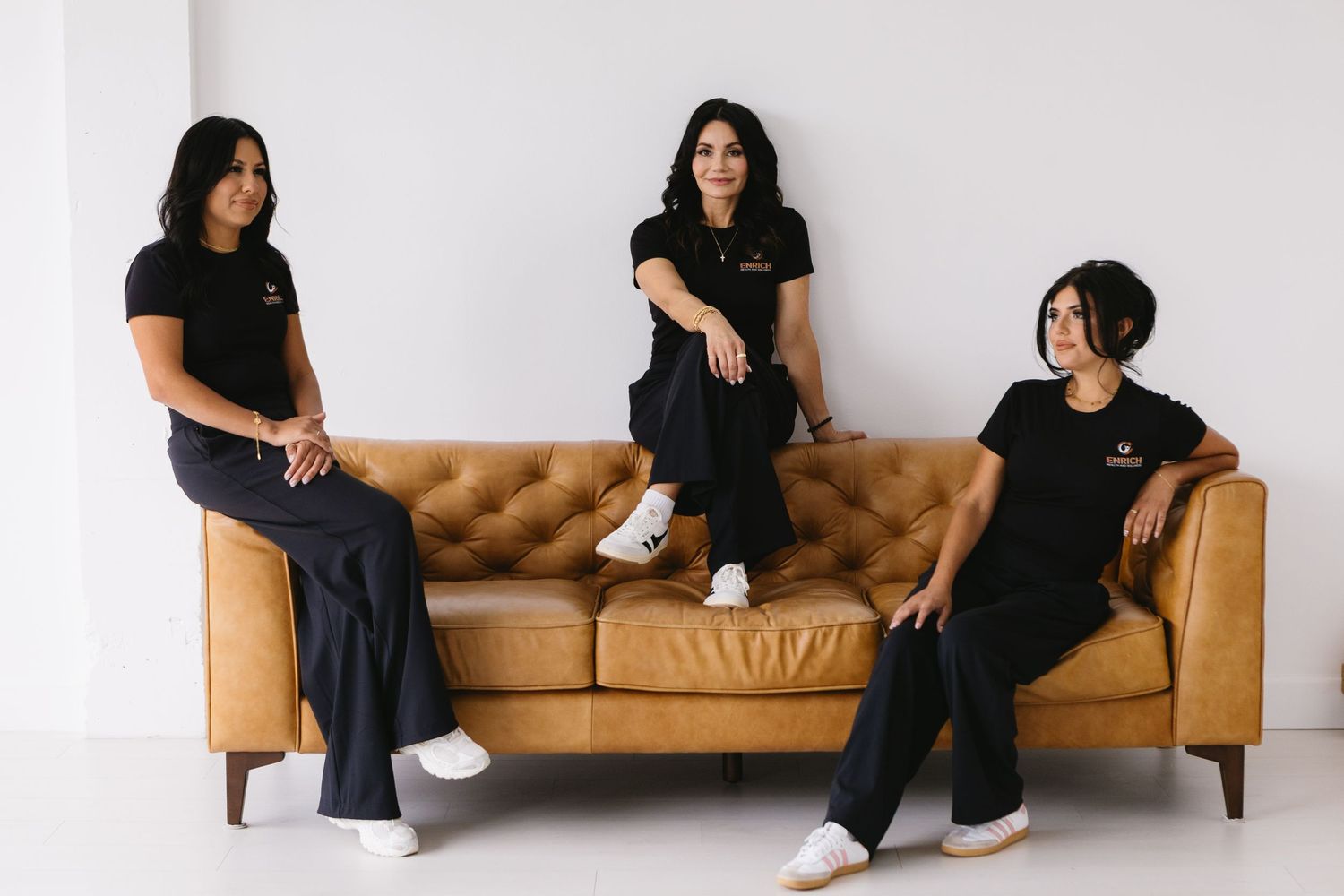 Three women in matching black uniforms pose on a tan leather sofa against a white background.