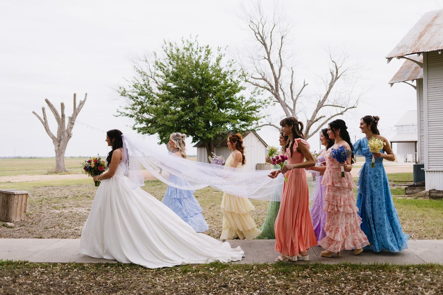 Bride in white gown with bridesmaids in colorful dresses holding bouquets outdoors at rustic farm wedding venue.