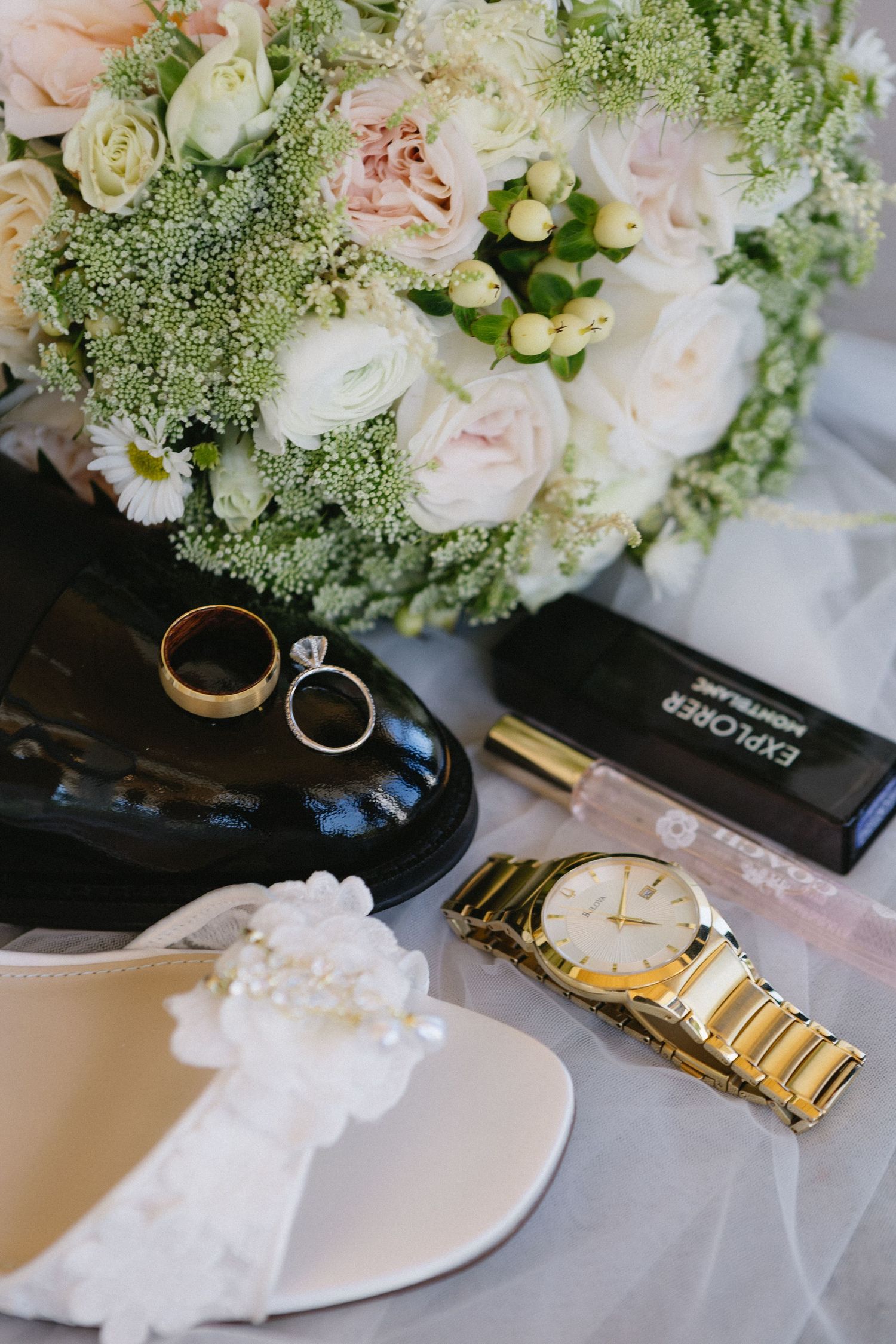 Wedding flat lay with bridal bouquet, gold rings, heels, watch, and perfume on white background.
