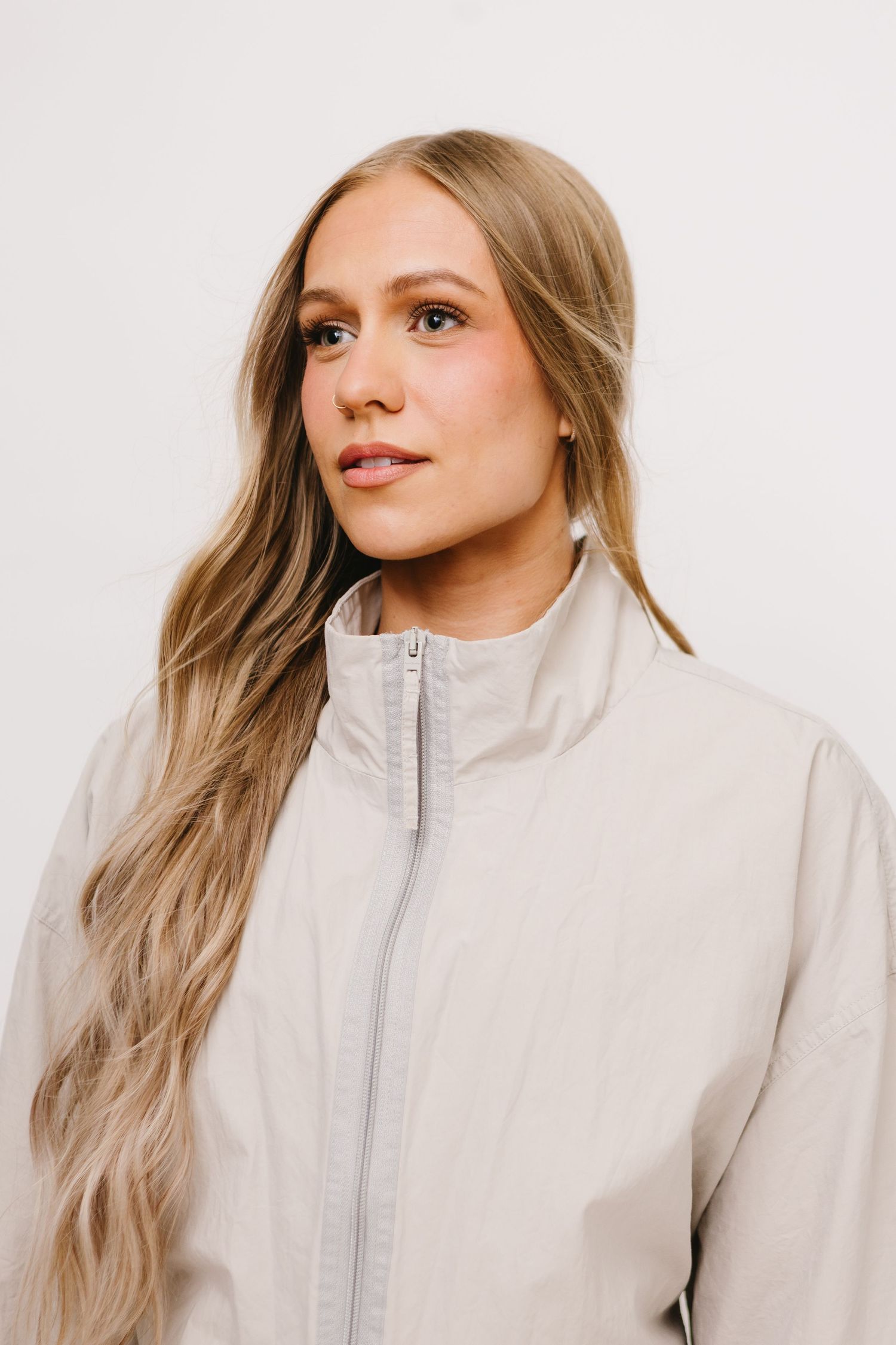 Young woman with long blonde hair wearing a beige zip-up jacket poses against a white background.