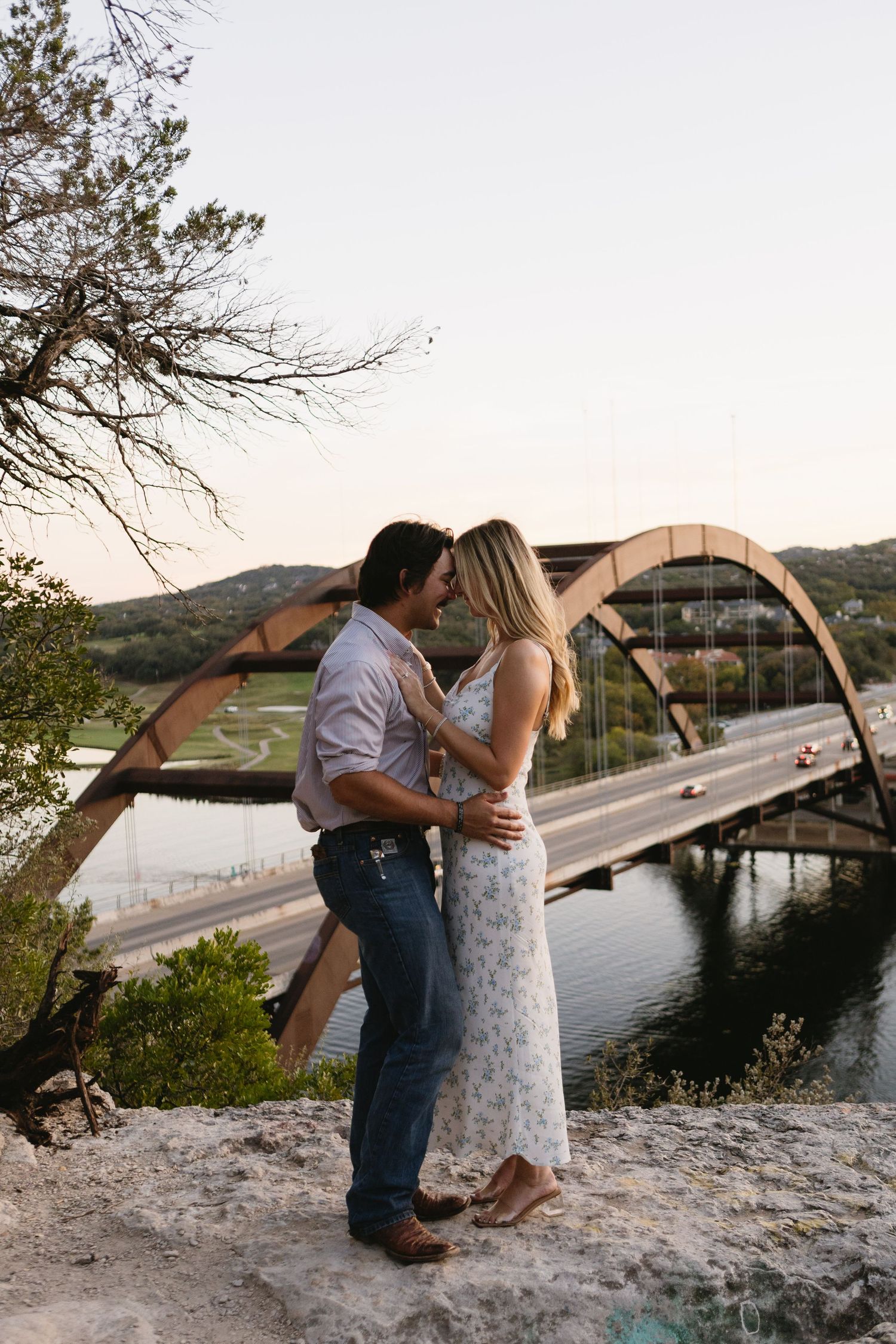 Couple embracing at sunset on rocky overlook with Pennybacker 360 Bridge and Colorado River in Austin, Texas background.