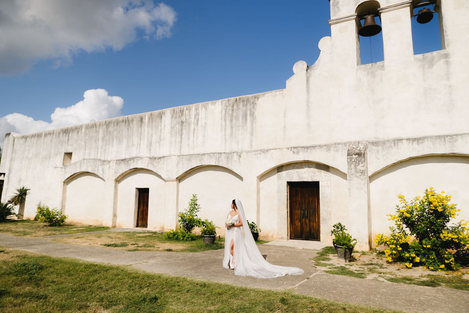 Bride in white gown walks outside a historic white stucco church with arched doorways under a blue sky.