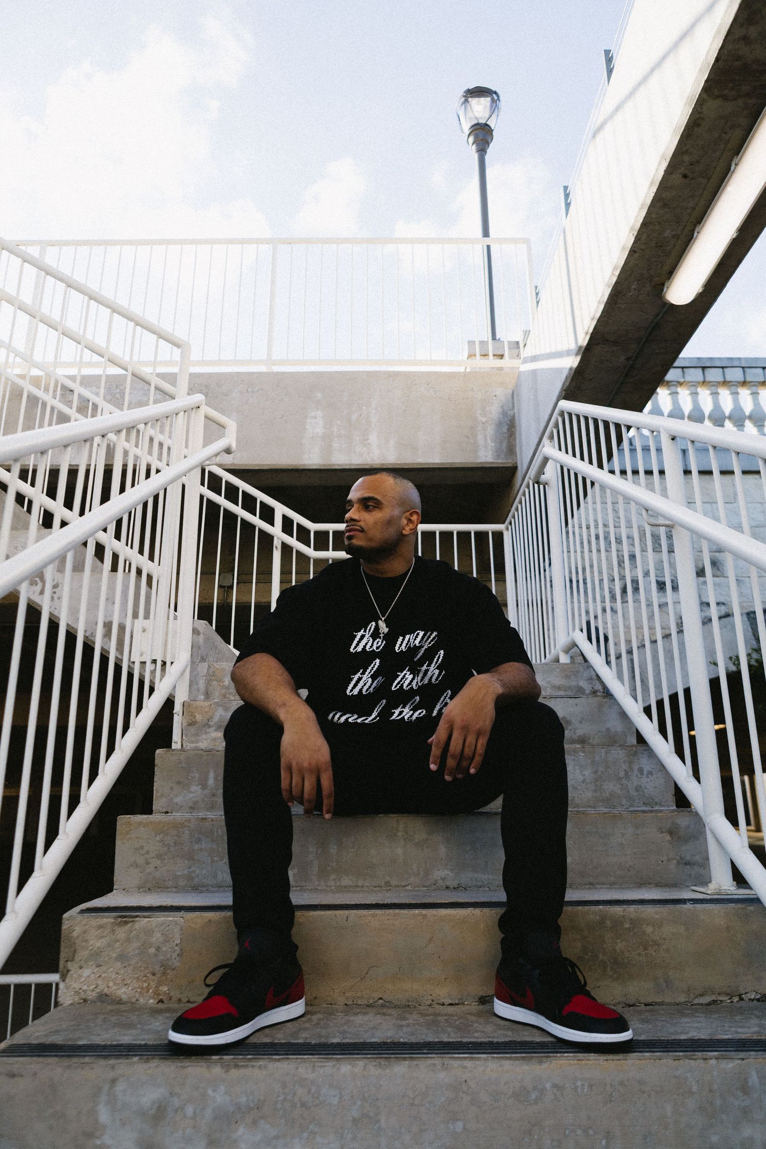 Man in black graphic tee and red sneakers sitting on concrete steps with white railings outdoors.