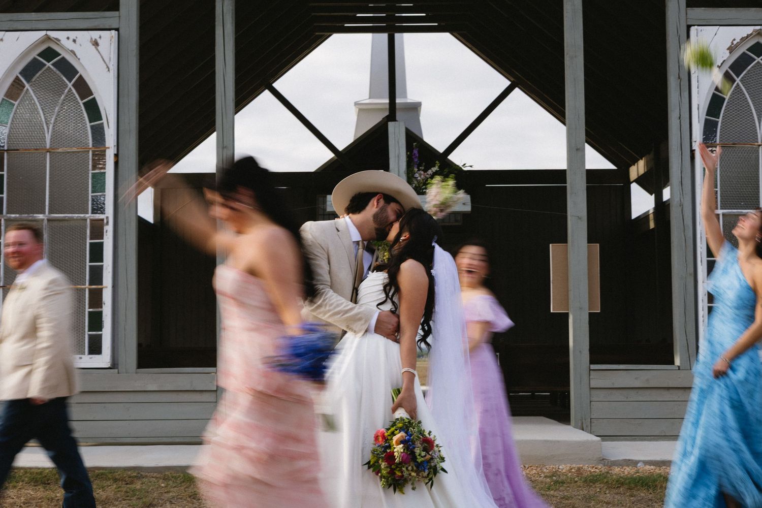 Bride and groom share a kiss outside a church, surrounded by wedding party members in colorful dresses.