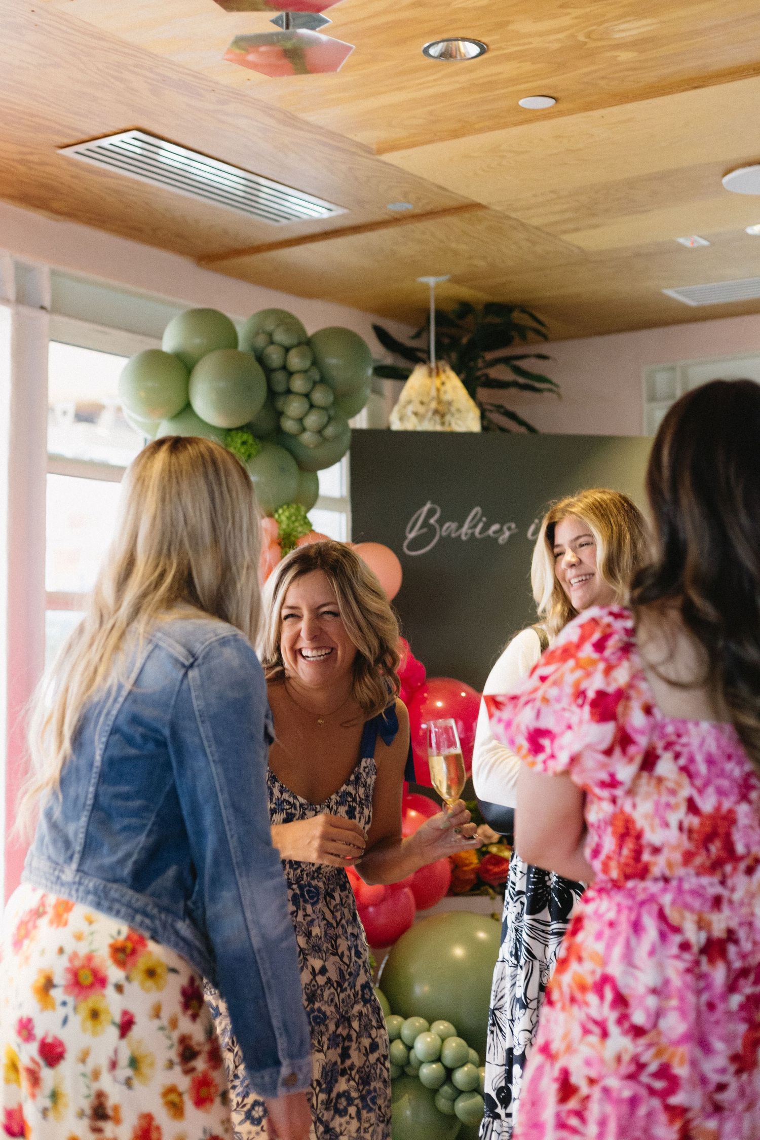 Women laughing and toasting with champagne at a baby shower with green and red balloon garland decorations.