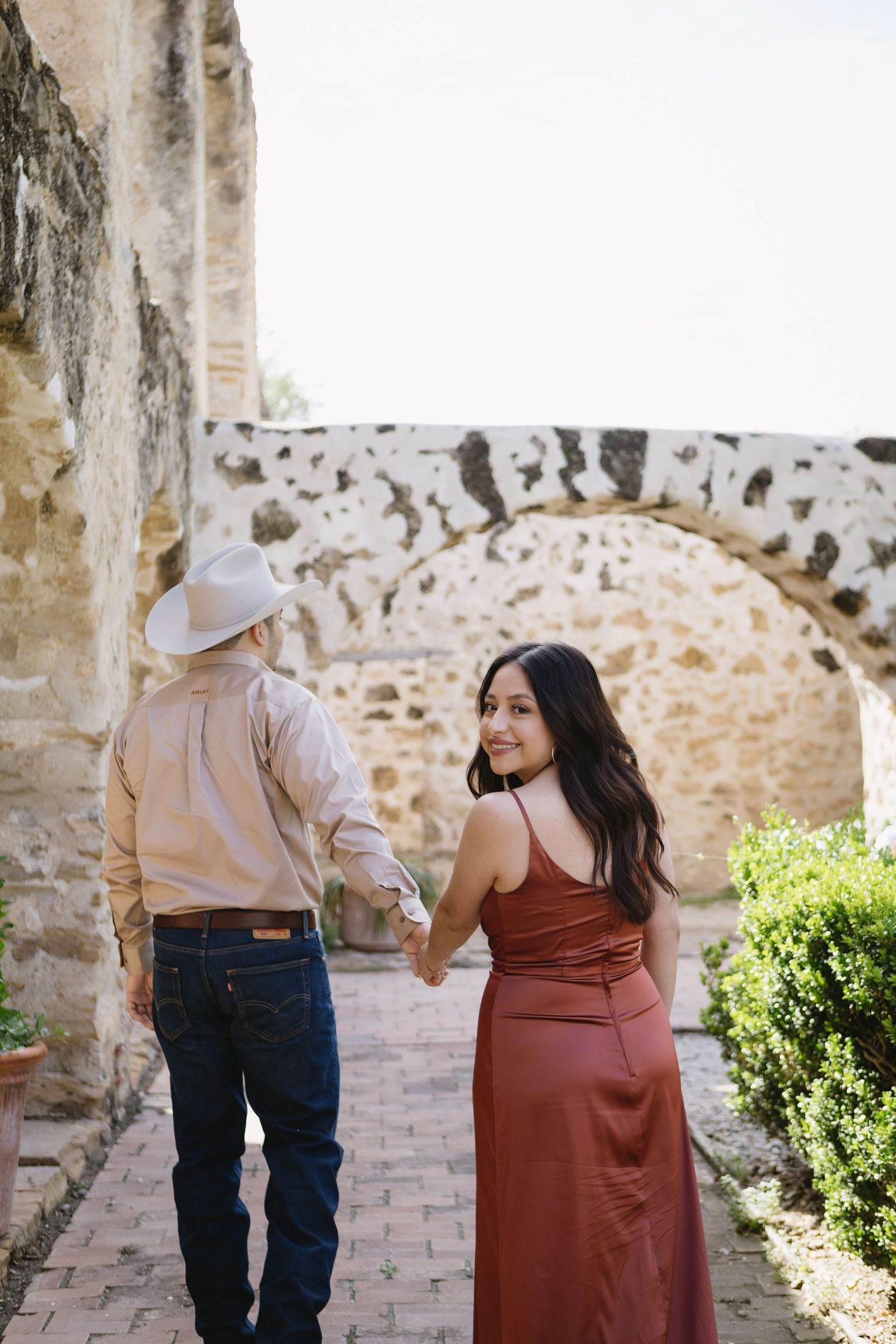 Couple holding hands at historic stone arch, woman in red satin dress looks back, man in cowboy hat and jeans.
