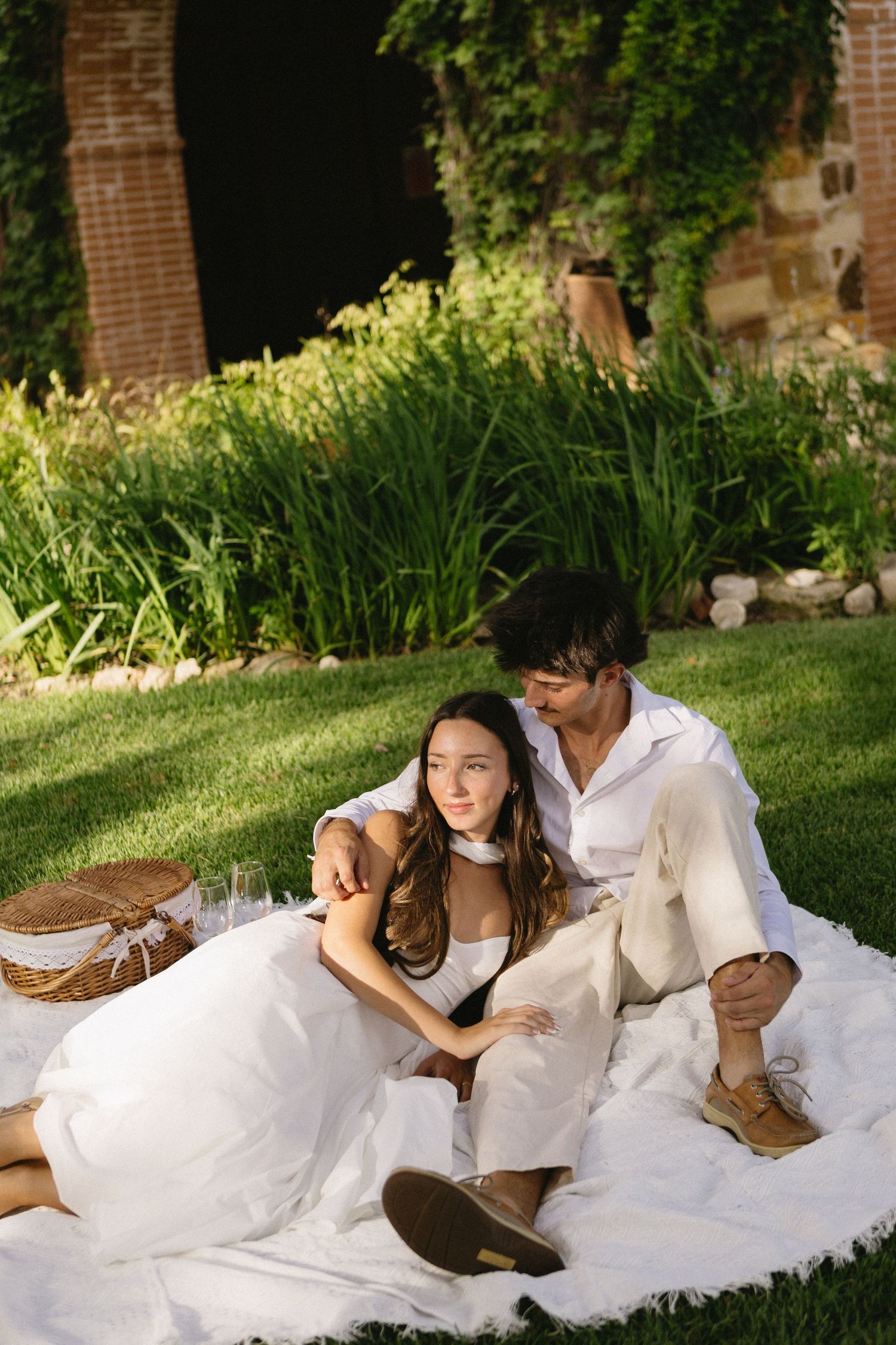 Couple in white attire enjoying a romantic garden picnic on a blanket with a wicker basket surrounded by lush greenery.