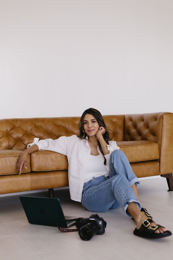 A person lounges beside a brown leather sofa wearing denim jeans and casual sandals.