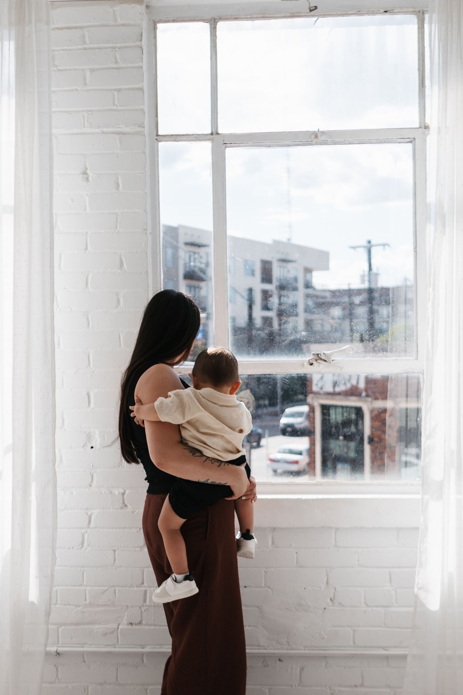 Parent in brown dress holding small child while standing in bright white room with large windows.