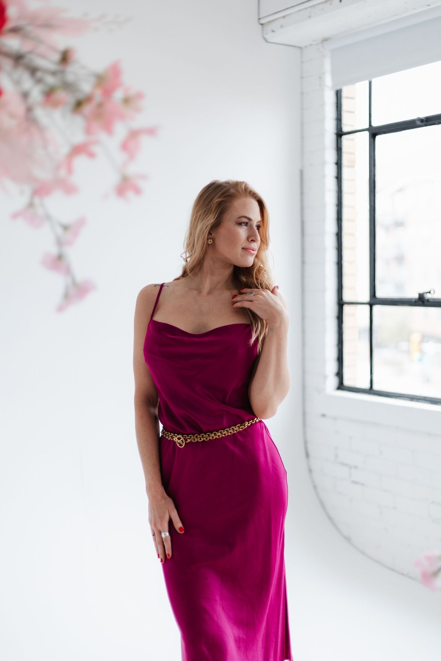 Person wearing magenta slip dress with chain belt poses in bright white studio.