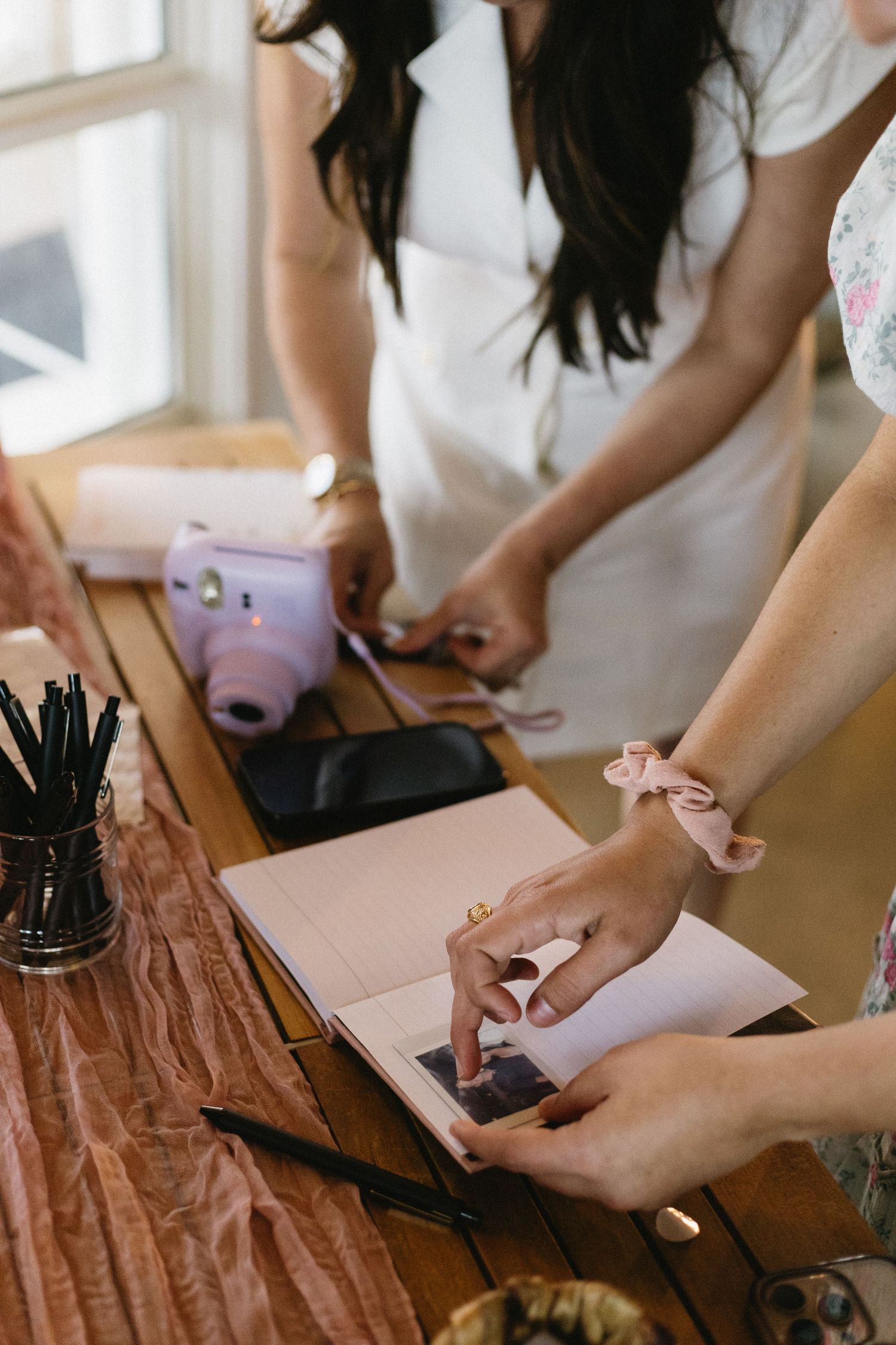 Two women arranging Instax photos in a pink album at a wooden table with a purple Fujifilm camera nearby.