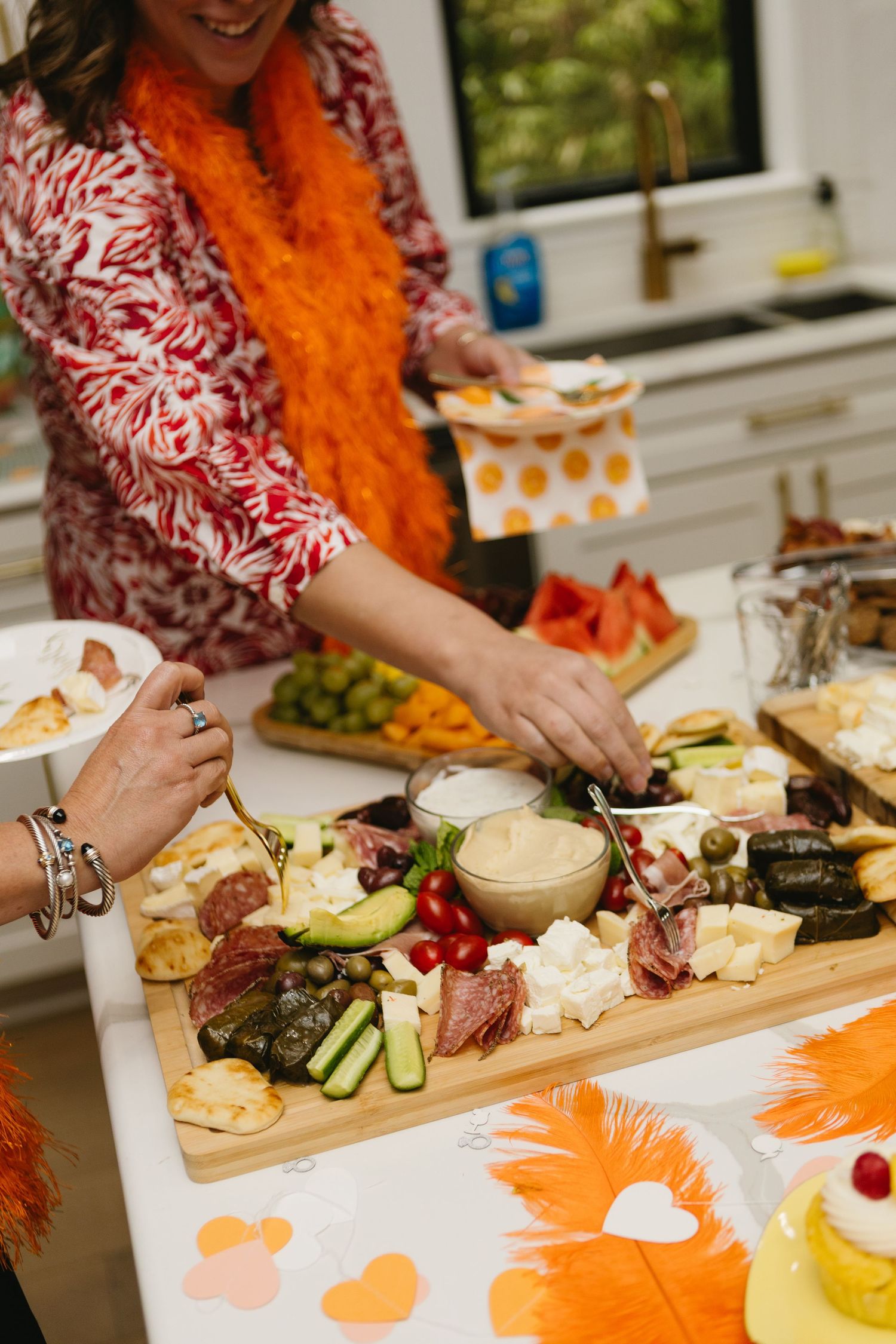 Woman in floral dress with orange boa reaches over charcuterie board at festive party spread with cheese, meats, and vegetables.