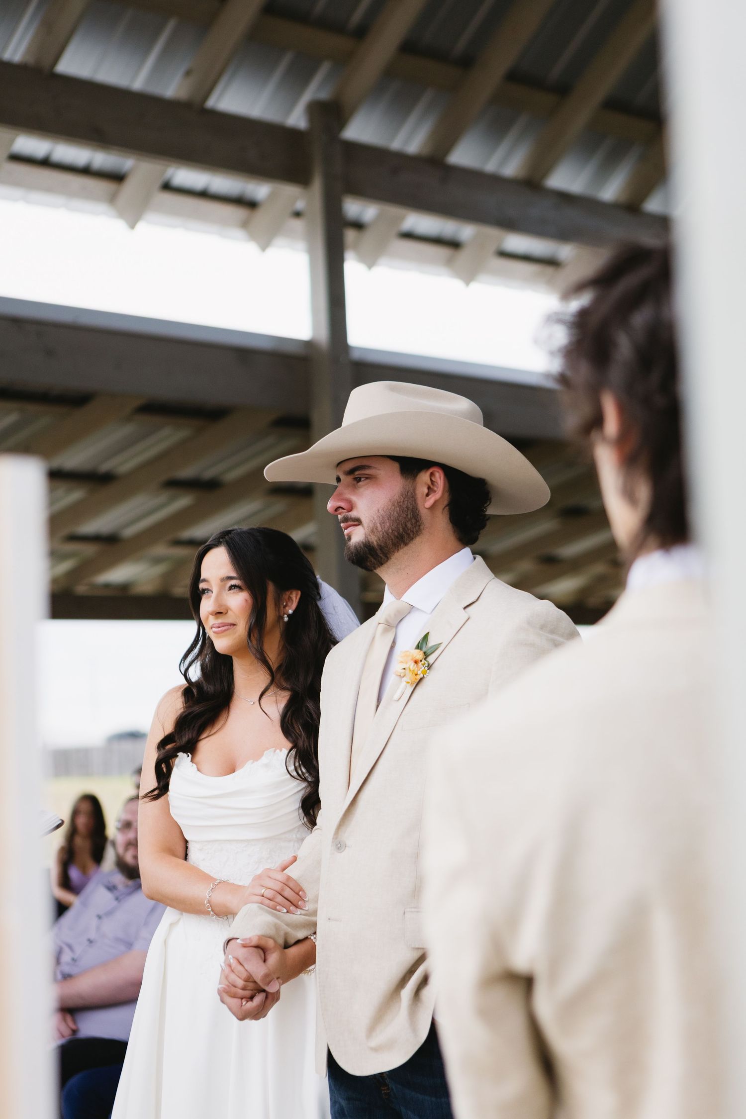 Bride and groom hold hands at outdoor wedding ceremony, groom wearing tan cowboy hat and suit with yellow boutonniere.