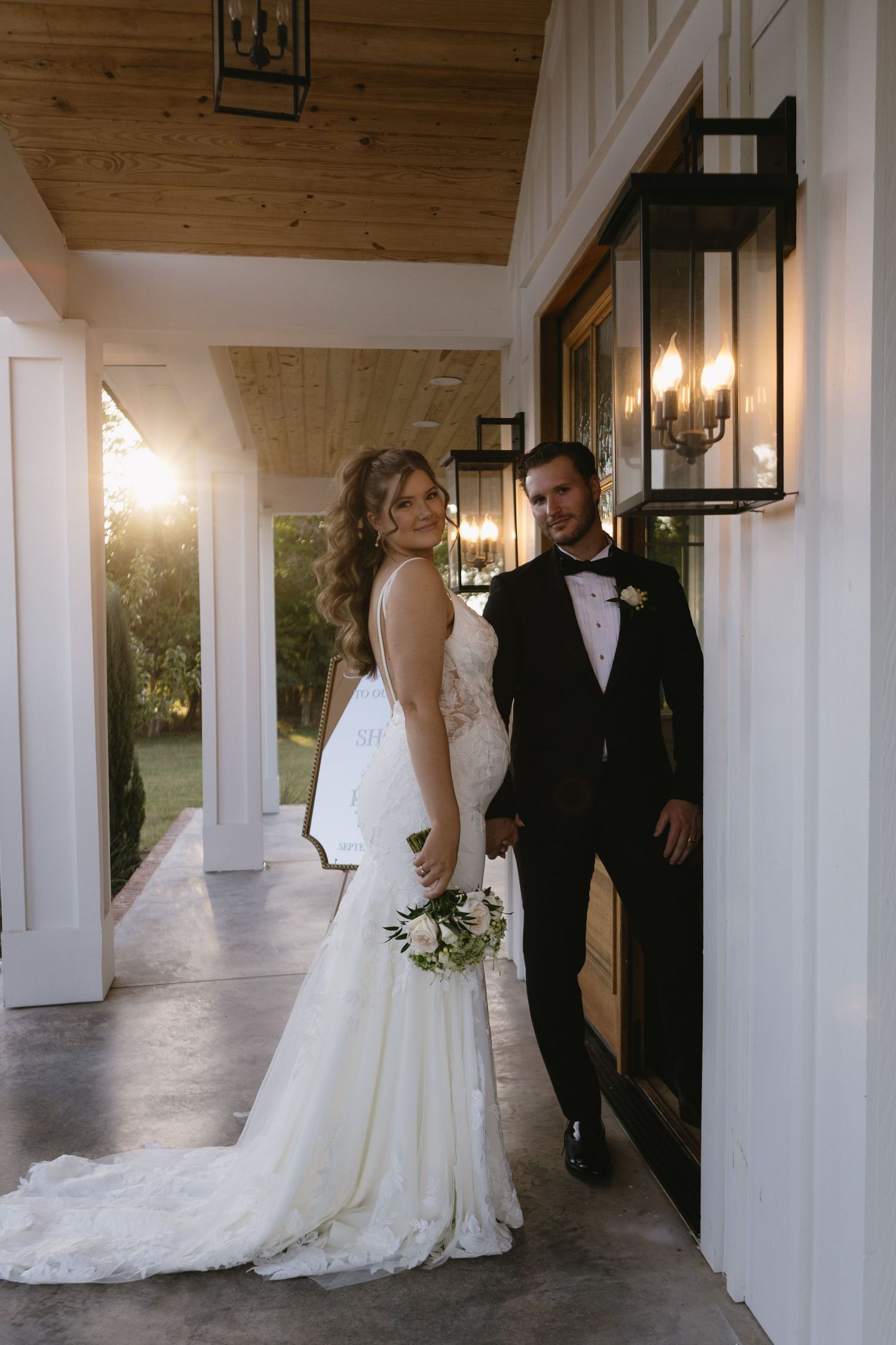 Bride in lace gown holding bouquet stands with groom in black tuxedo on elegant white venue porch at golden hour.