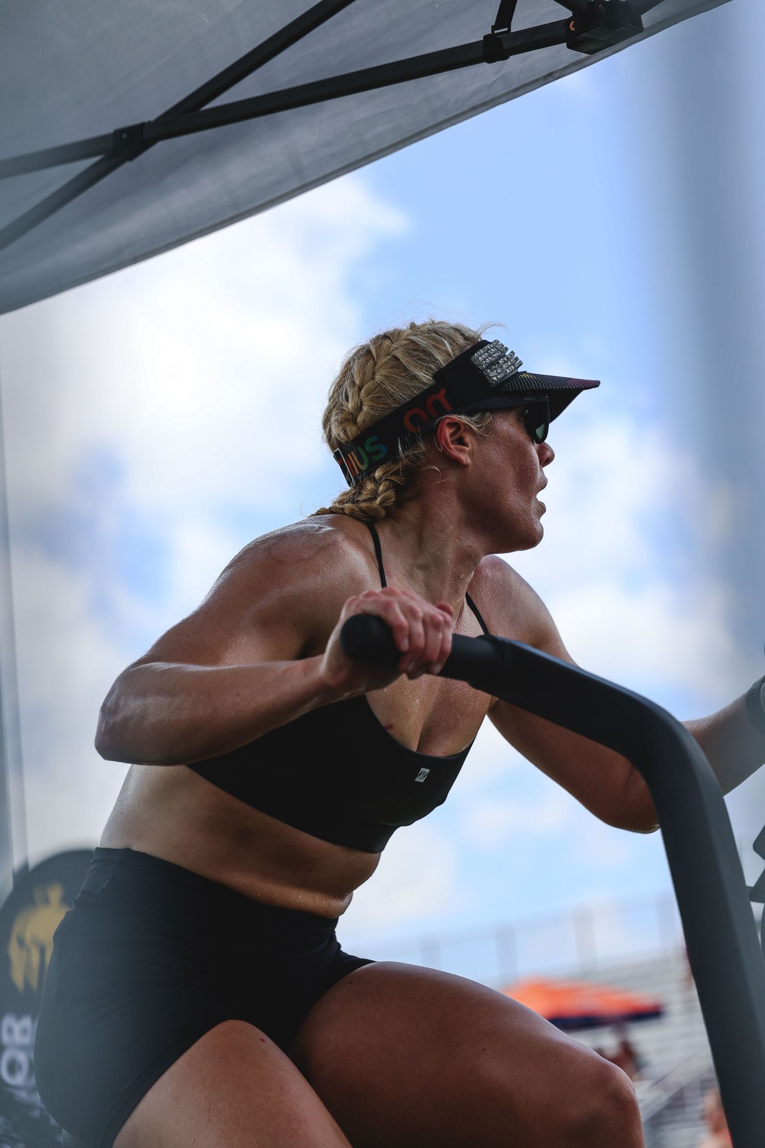 Athletic woman in sports bra and visor exercises on stationary bike outdoors under tent canopy on cloudy day.