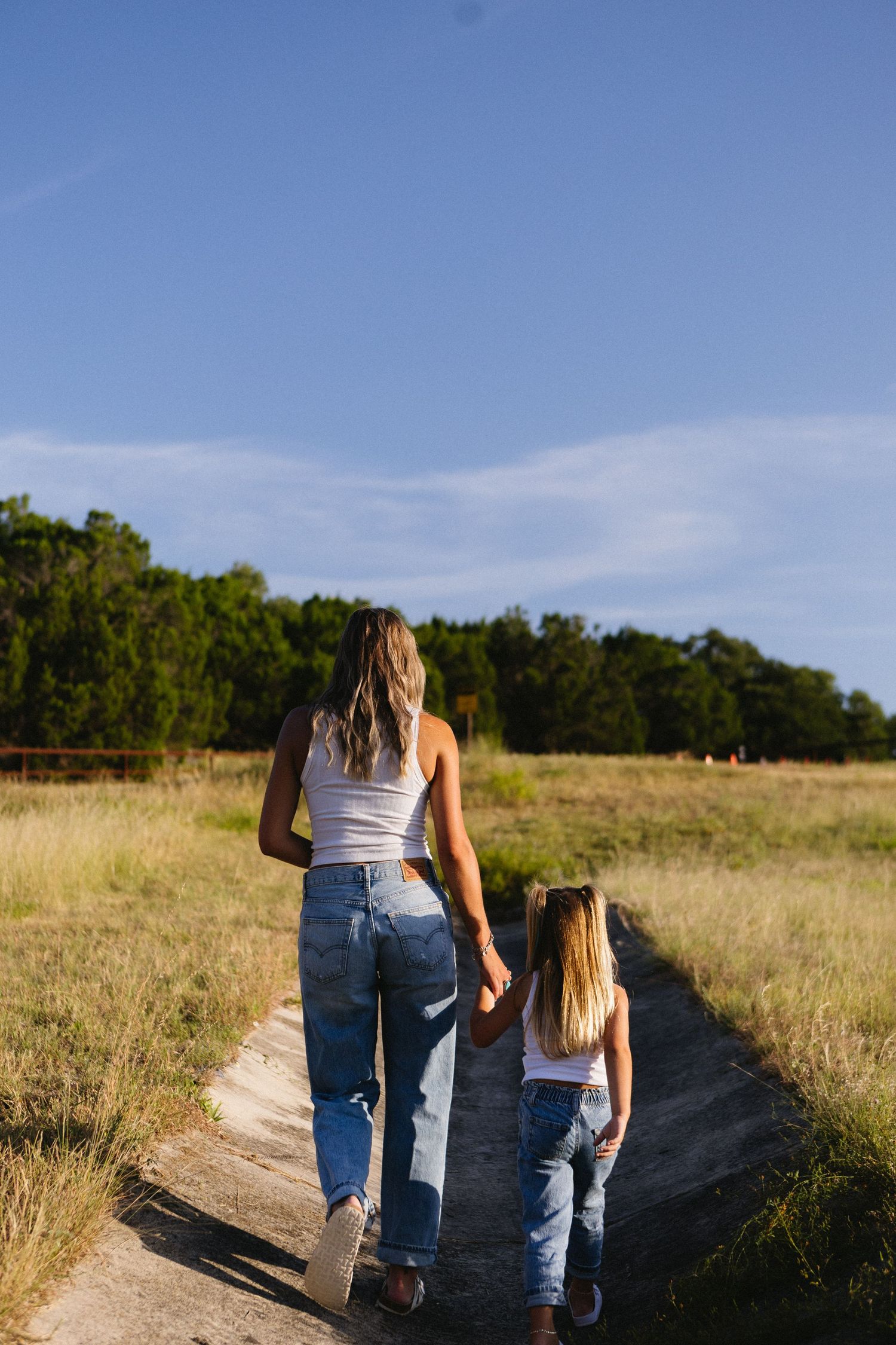 Mother and daughter in matching white tops and jeans walk hand-in-hand down a rural path surrounded by green fields.